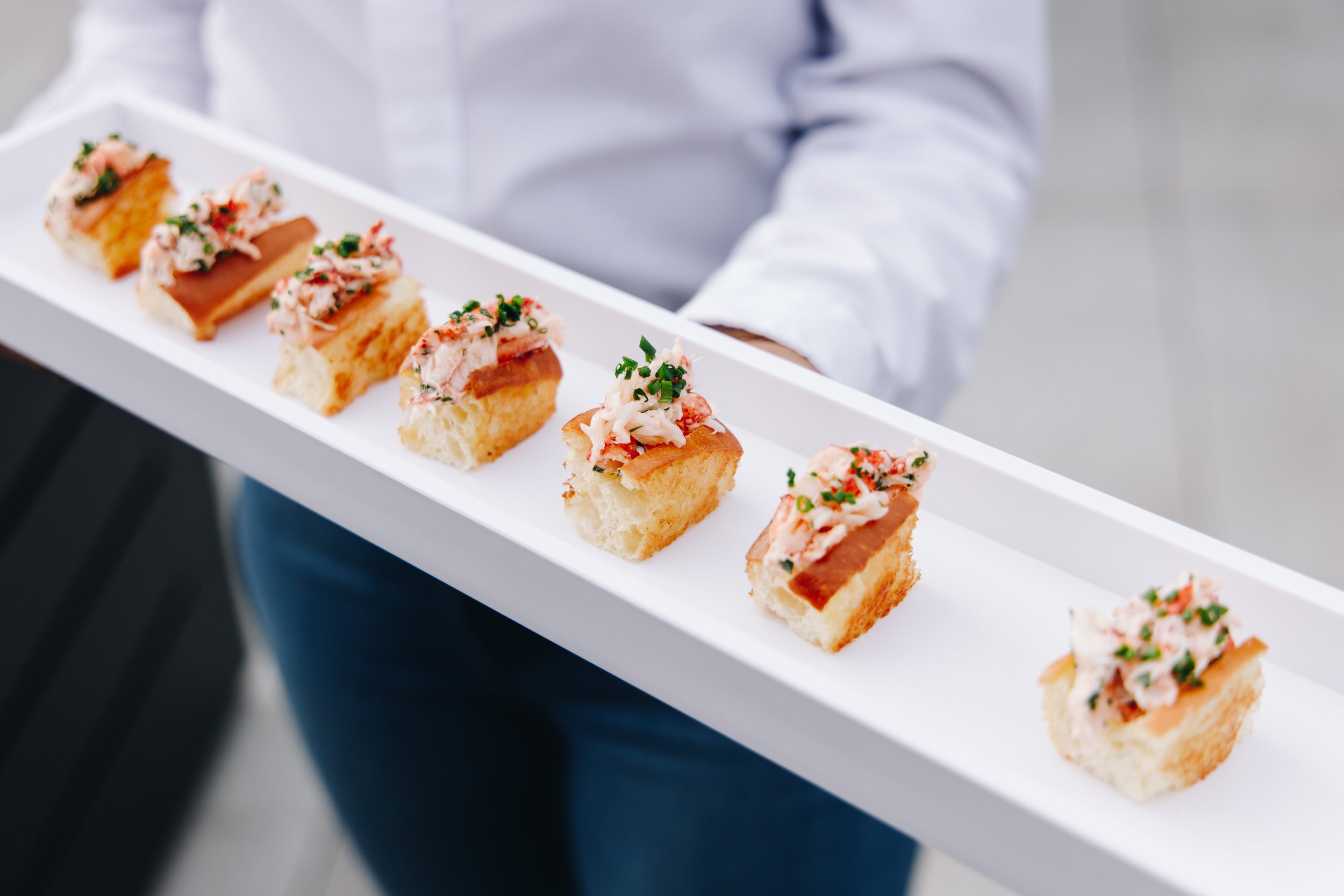A person in a white shirt holding a white tray with seven pieces of canapé topped with seafood and herbs.