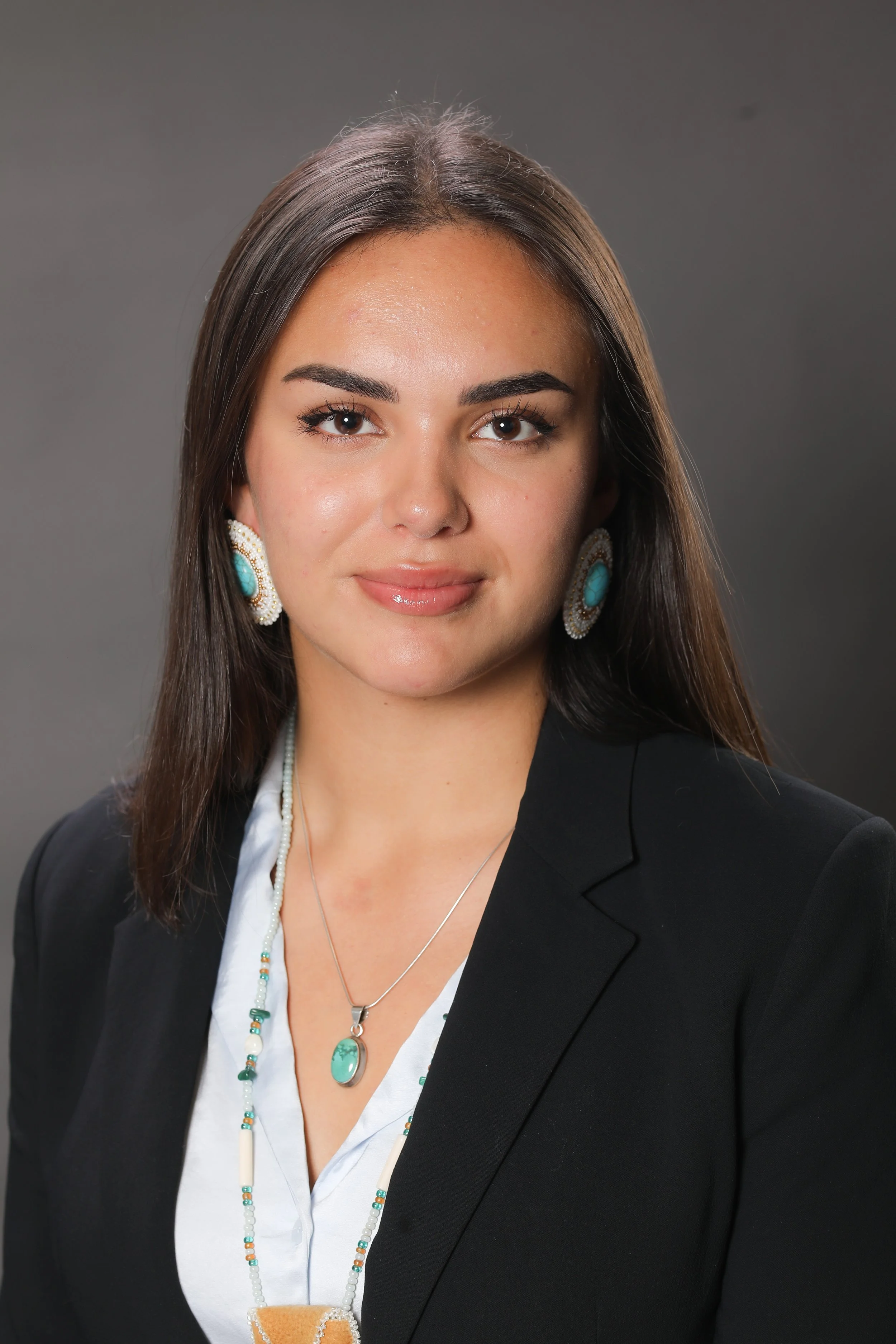 A woman with long brown hair, wearing a black blazer, white shirt, and turquoise jewelry, posing against a gray background.