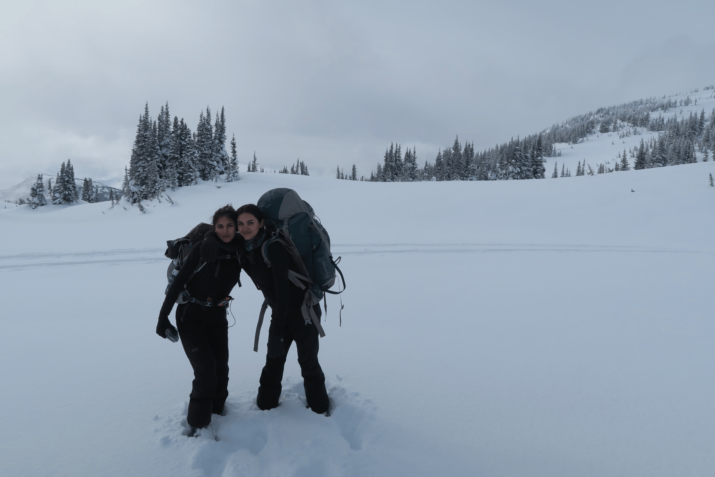 Two women in black winter clothing and backpacks pose together on snow in a snowy mountain landscape with pine trees and overcast sky.