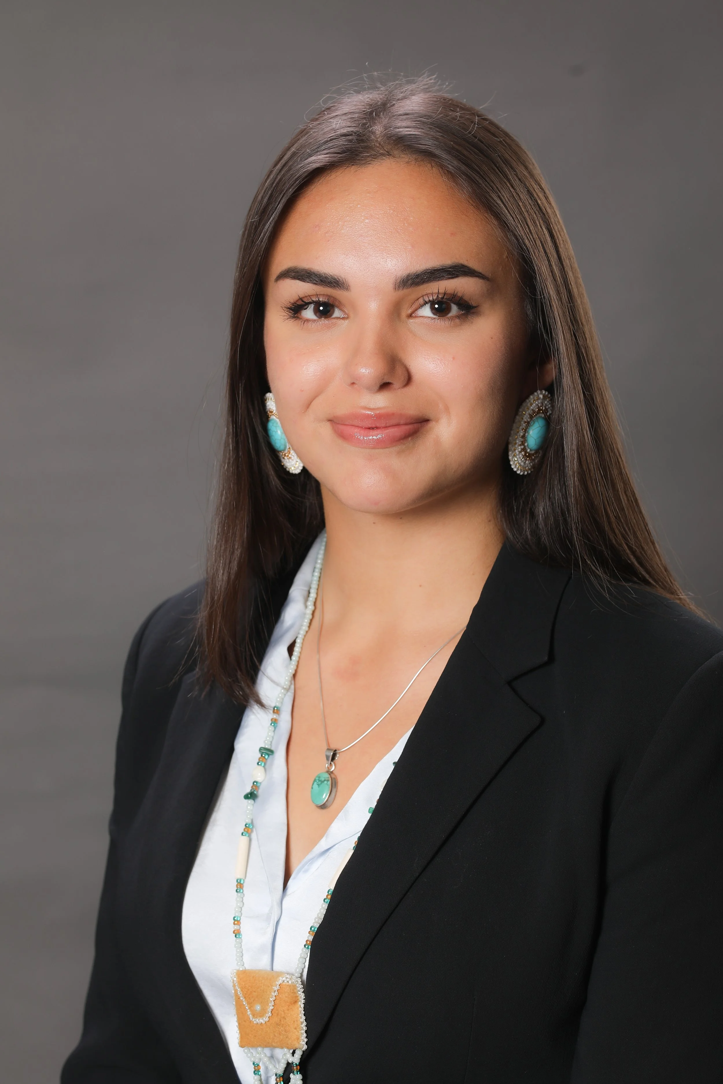 Portrait of a young woman with long brown hair wearing a black blazer, white shirt, turquoise earrings, and matching turquoise jewelry, standing against a plain gray background.