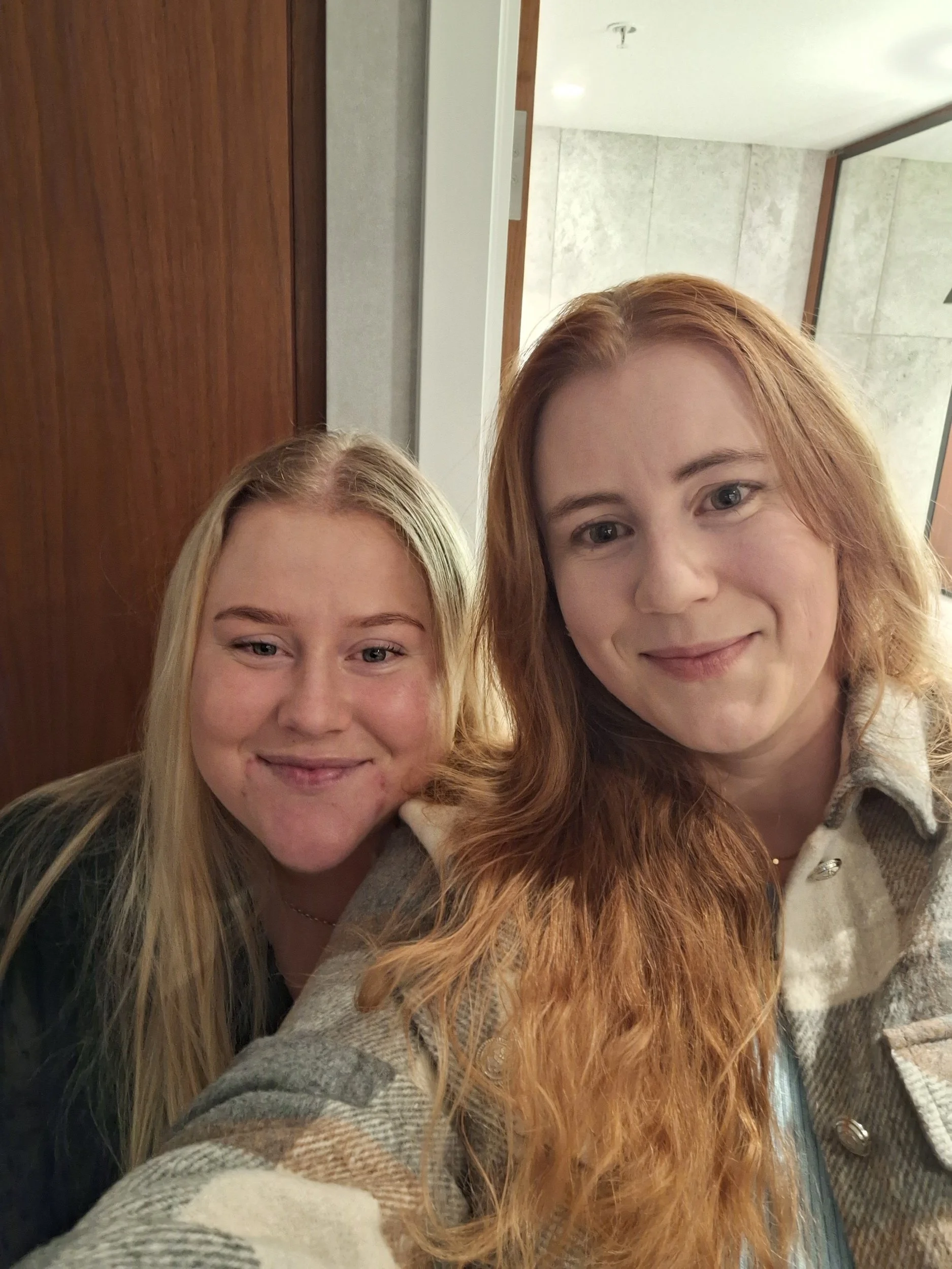 Two smiling women taking a selfie together indoors, with part of their faces and hair visible.