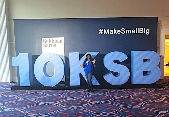 A woman standing in front of large 3D letters spelling '$10K SB' at a Goldman Sachs event, with a sign '#MakeSmallBig' on a large screen behind her.