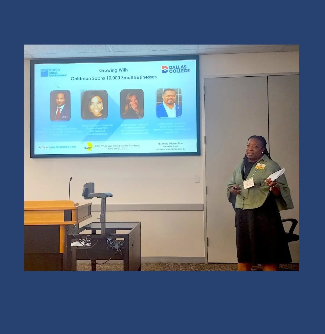 A woman giving a presentation in a classroom with a large screen displaying information about small businesses and a panel of four speakers, in a university setting.