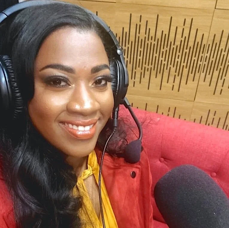 Woman with dark hair and makeup wearing a yellow top, red jacket, and black headphones with a microphone, sitting on a red couch in a studio with wooden acoustic panels.