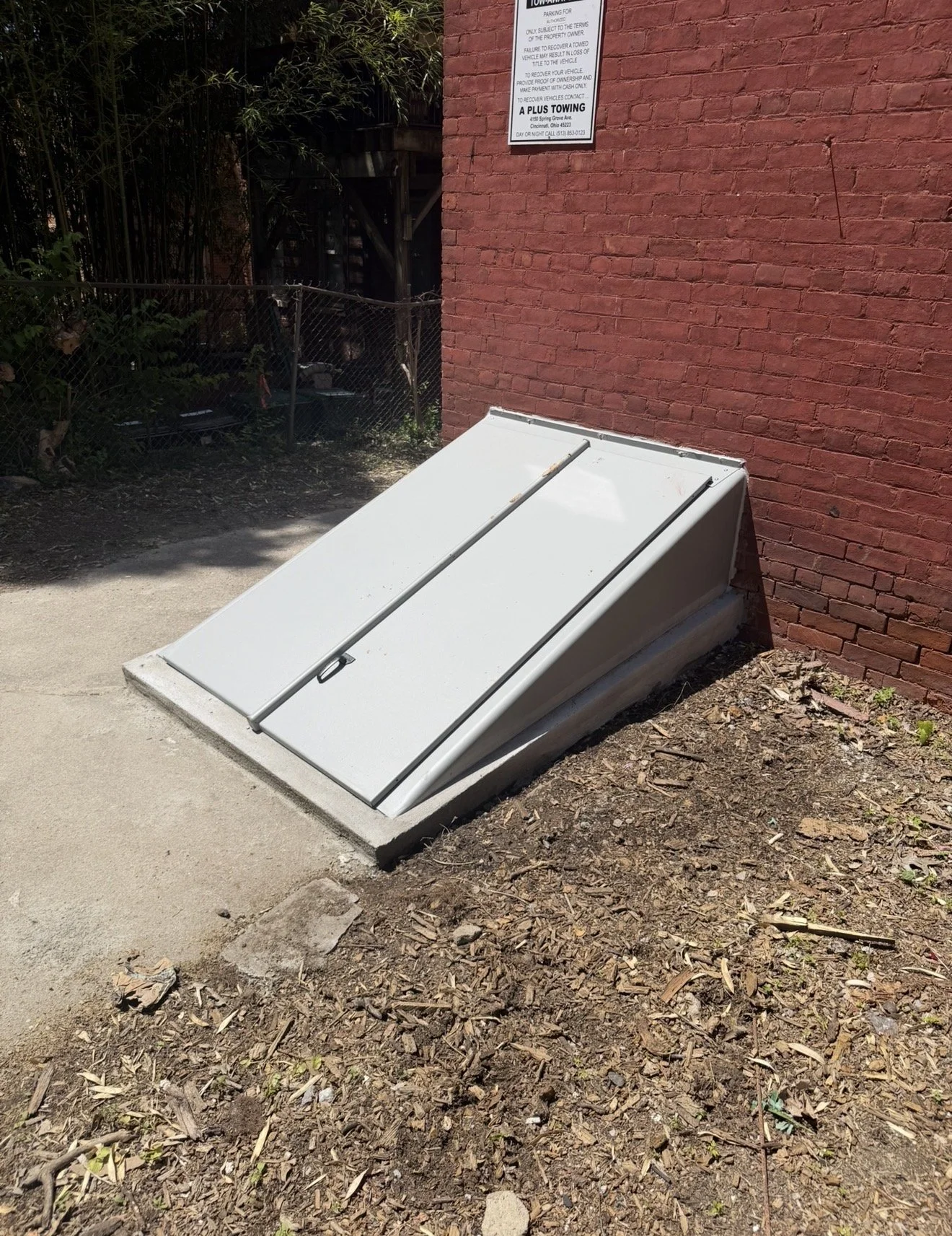 A handicap wheelchair lift installed at the base of a red brick building, with a white cover and a handle, on a patch of dirt and sidewalk.