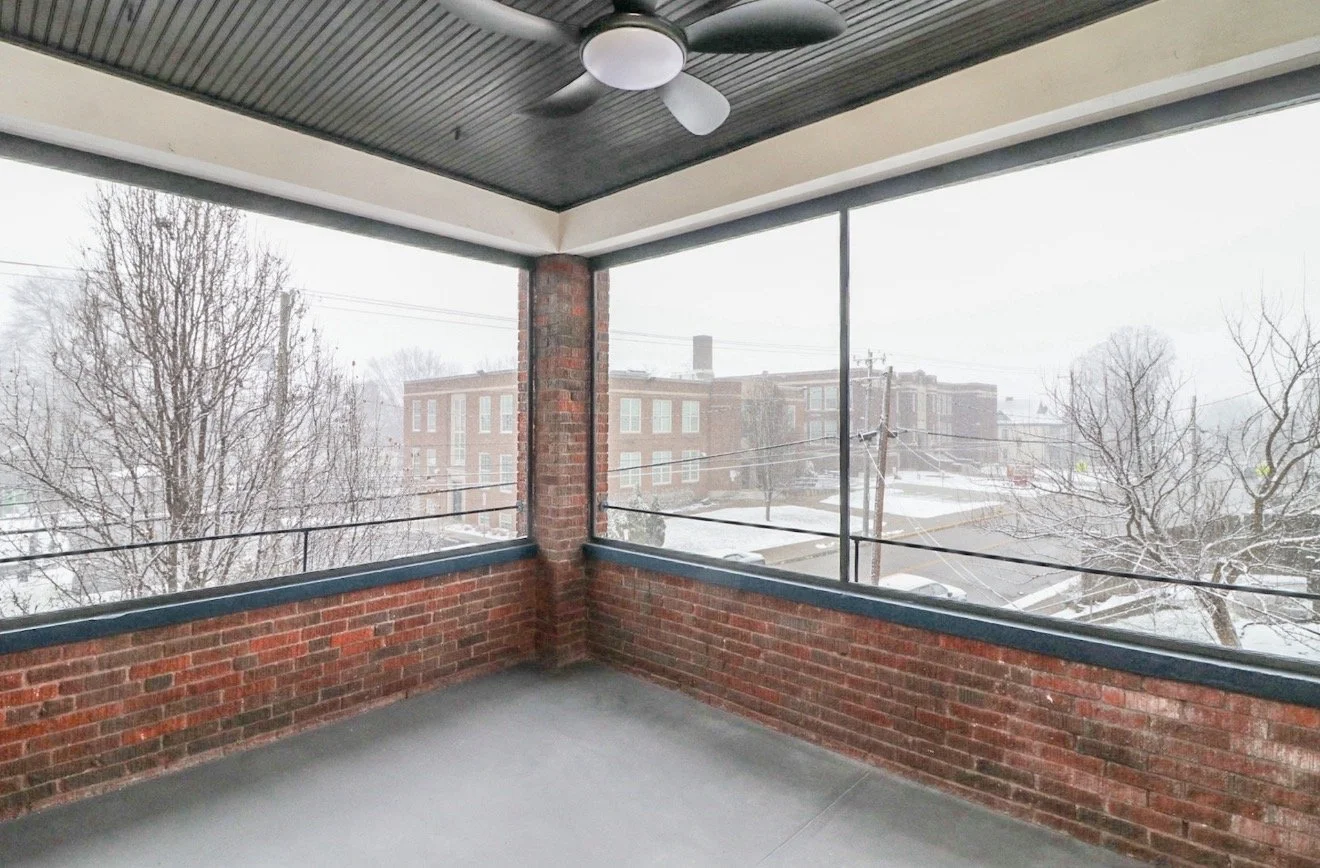 A covered outdoor balcony with brick walls, glass screens, a ceiling fan, and a snowy view of trees and buildings outside.