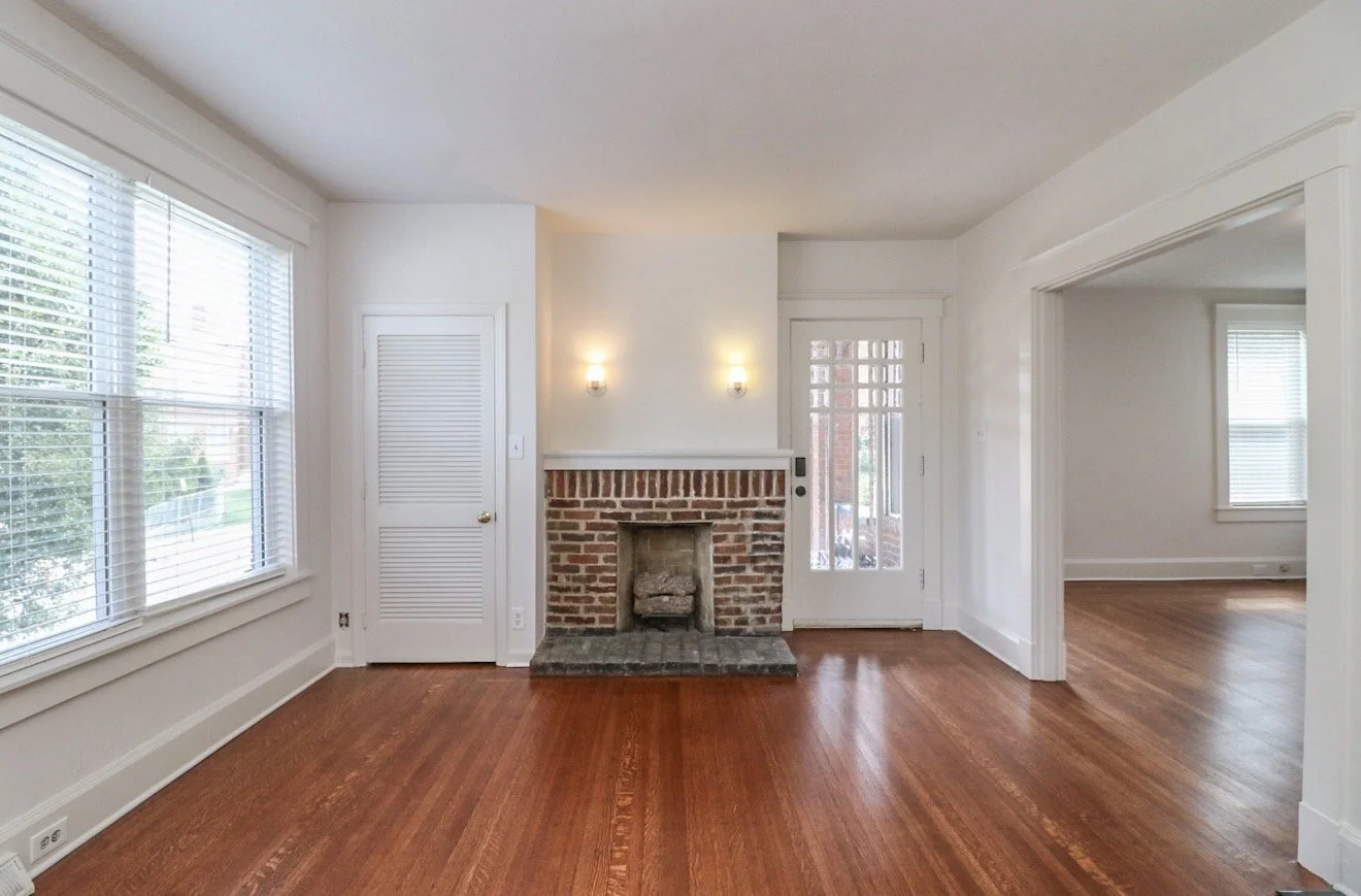 Empty living room with hardwood floors, a brick fireplace, large windows with white blinds, and a door leading outside.