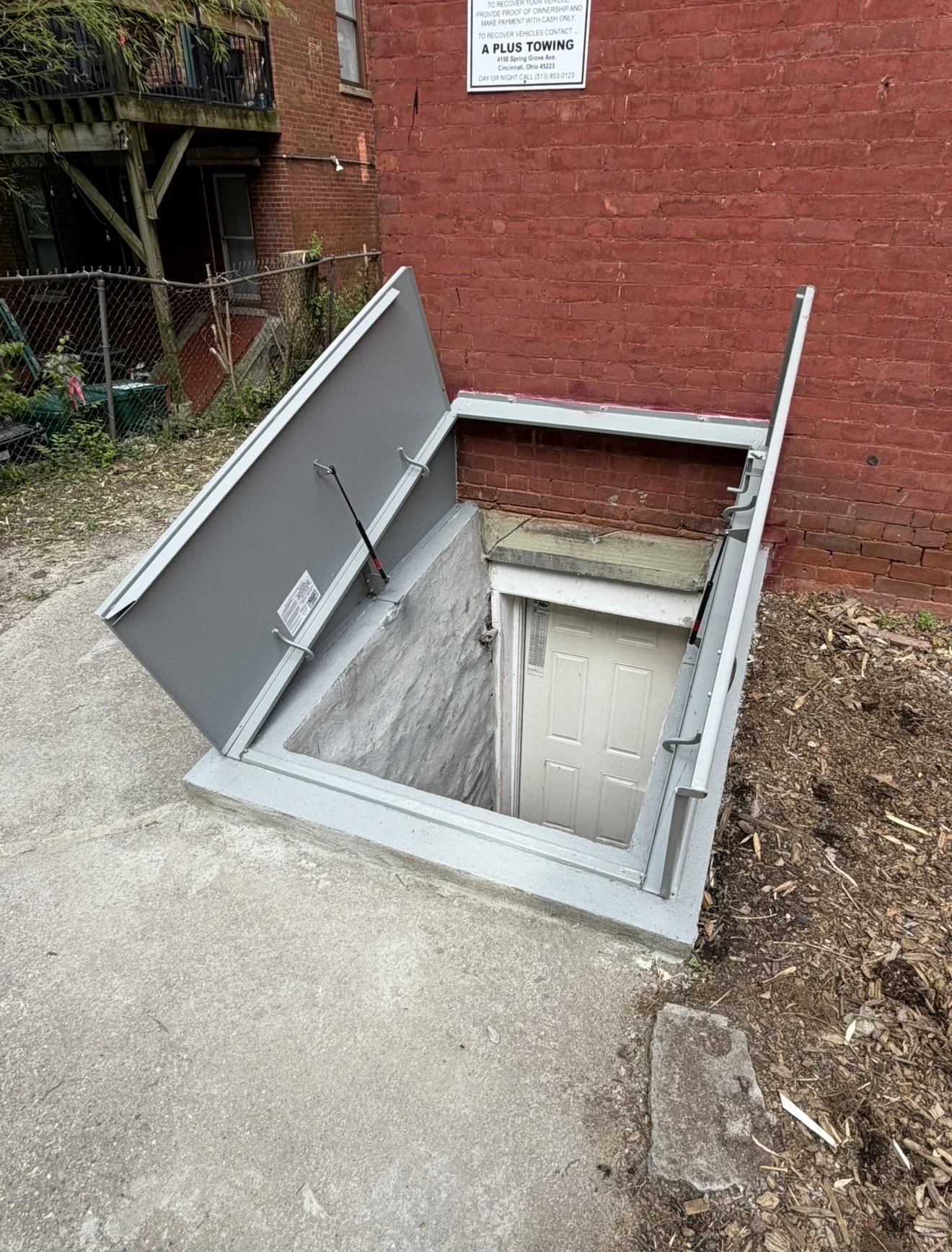 A rectangular underground basement entrance with a metal cover propped open, revealing a descending staircase with a white door at the bottom. The entrance is on a concrete sidewalk next to a red brick building, with surrounding dirt and soil on the 