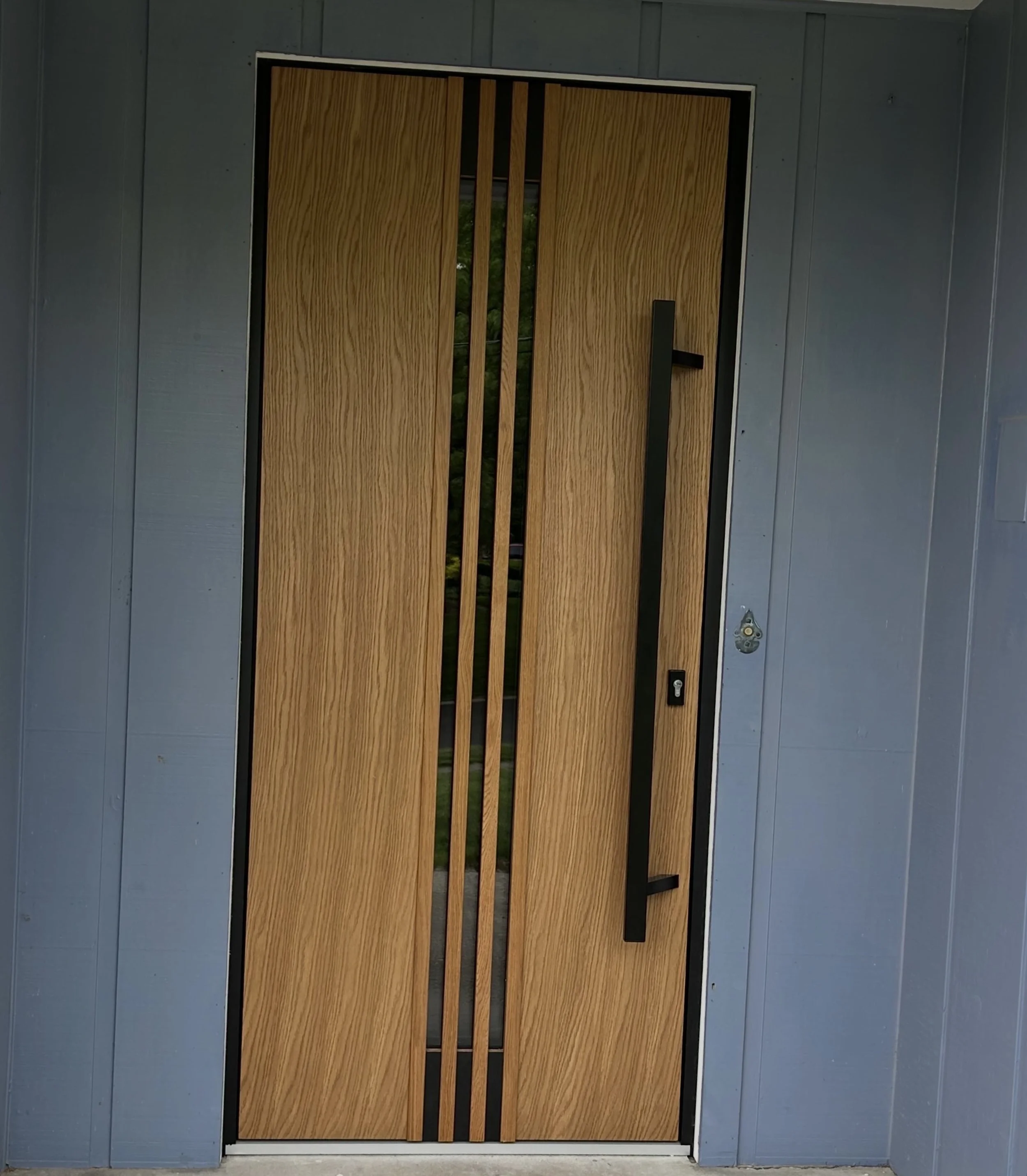 Modern wooden door with vertical glass panels and black metal frame, installed in a blue exterior wall.