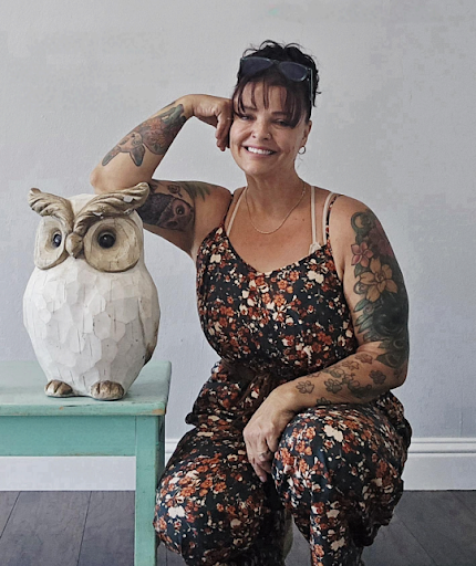 Woman with tattoos sitting next to a large owl sculpture on a table.
