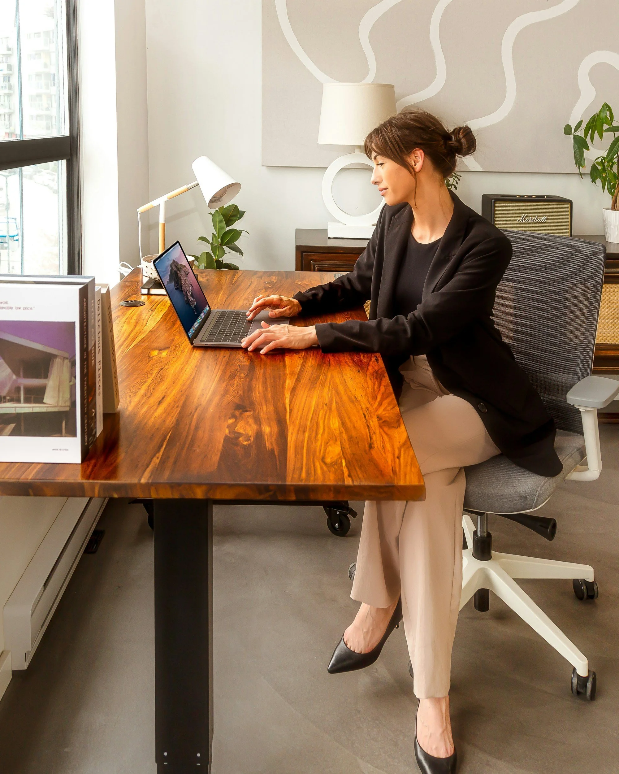 Woman in black blazer and beige pants sitting at wooden desk, working on laptop, in modern office with window, desk lamp, plants, and audio speaker.