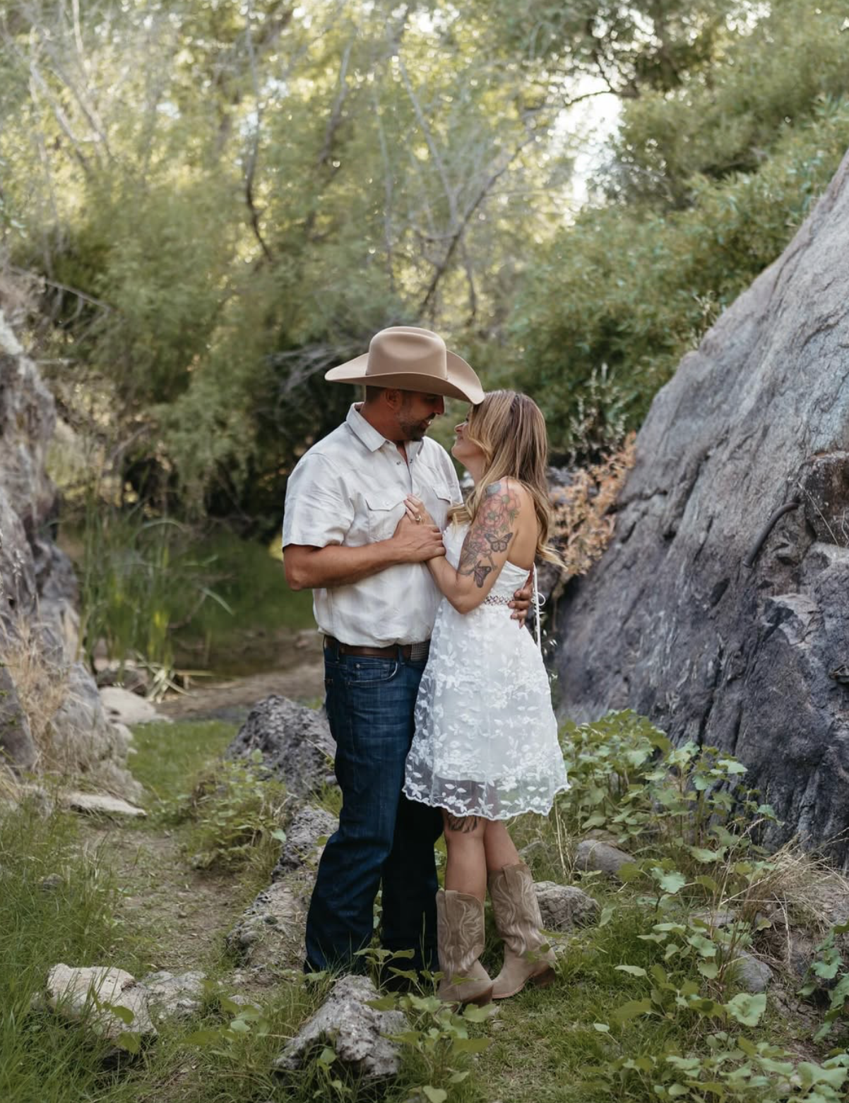 A couple standing on a grassy path surrounded by rocks and trees, sharing an intimate moment. The man wears a cowboy hat, white shirt, and jeans, while the woman wears a white dress with lace details and cowboy boots.