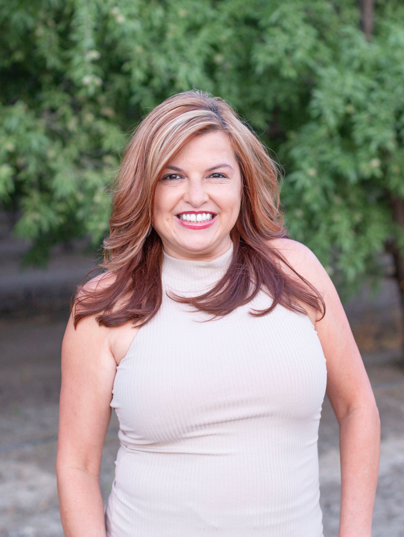 A woman with shoulder-length wavy reddish-brown hair smiling outdoors with green leafy trees in the background, wearing a sleeveless beige turtleneck top.