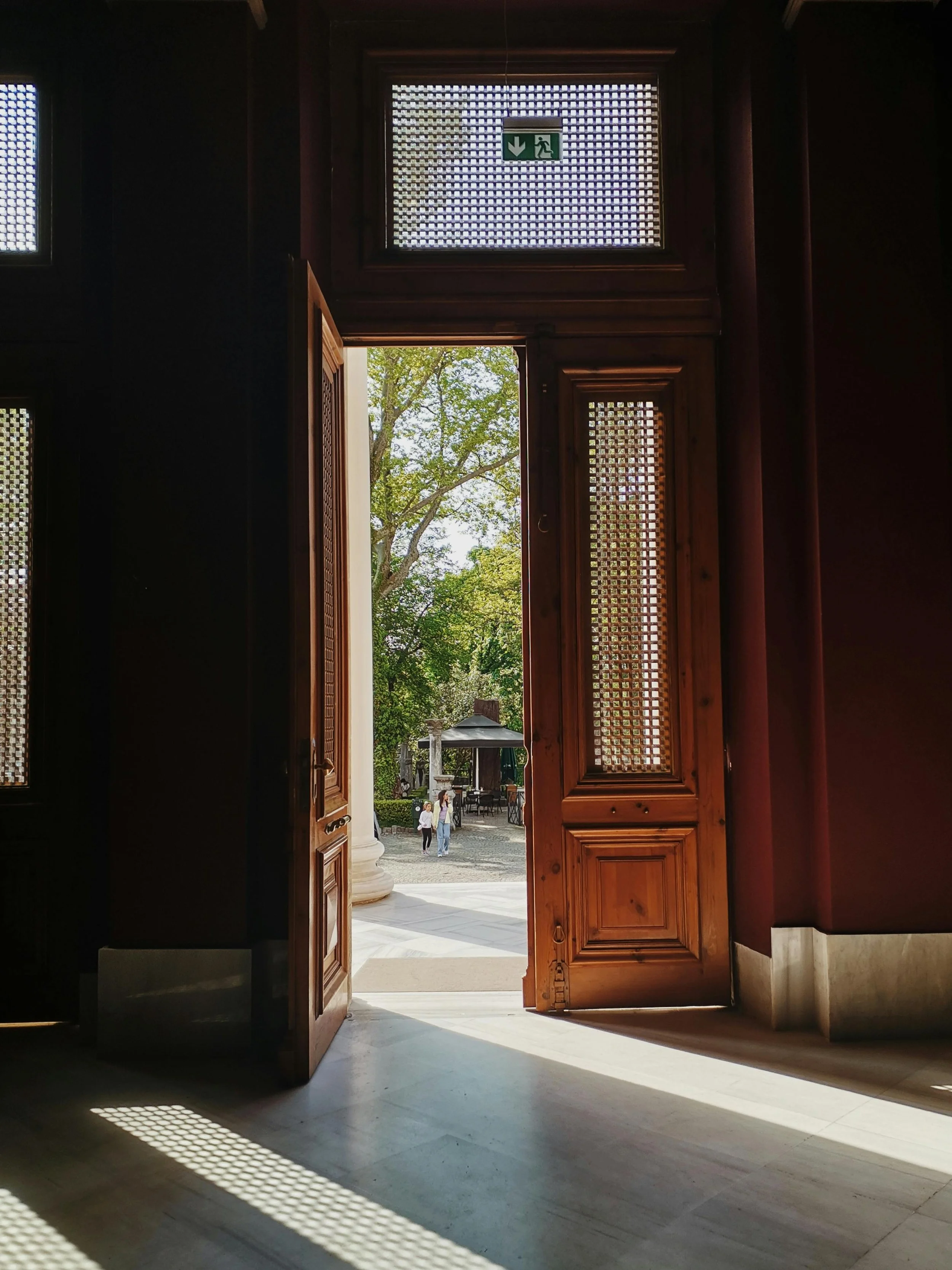 Open wooden door leading outside to a courtyard with trees and a gazebo, sunlight casting shadows inside.