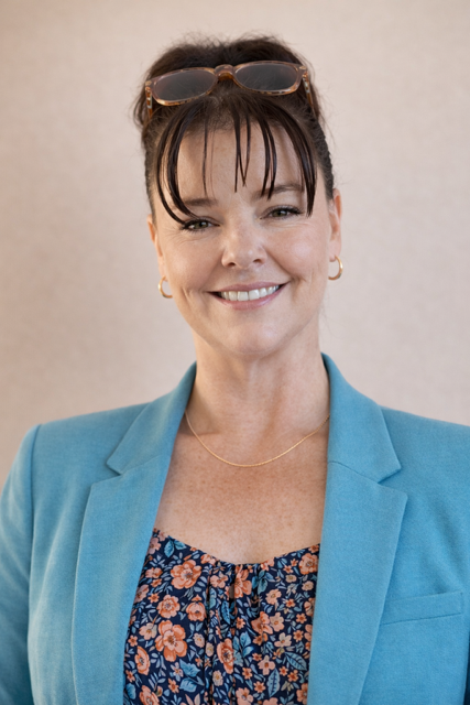 Lesley Curry, A woman smiling, wearing sunglasses on her head, gold hoop earrings, a thin gold necklace, a blue blazer, and a floral top, standing against a plain background.