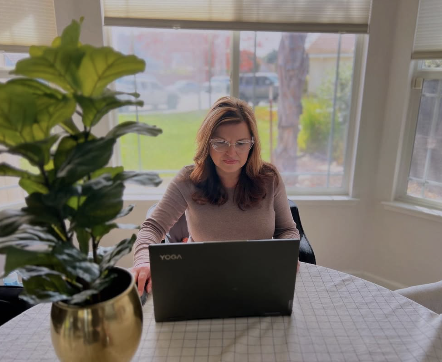 A woman with glasses working on a laptop at a round table, with a large potted green plant in the foreground and a window with a view of a lawn and trees outside.