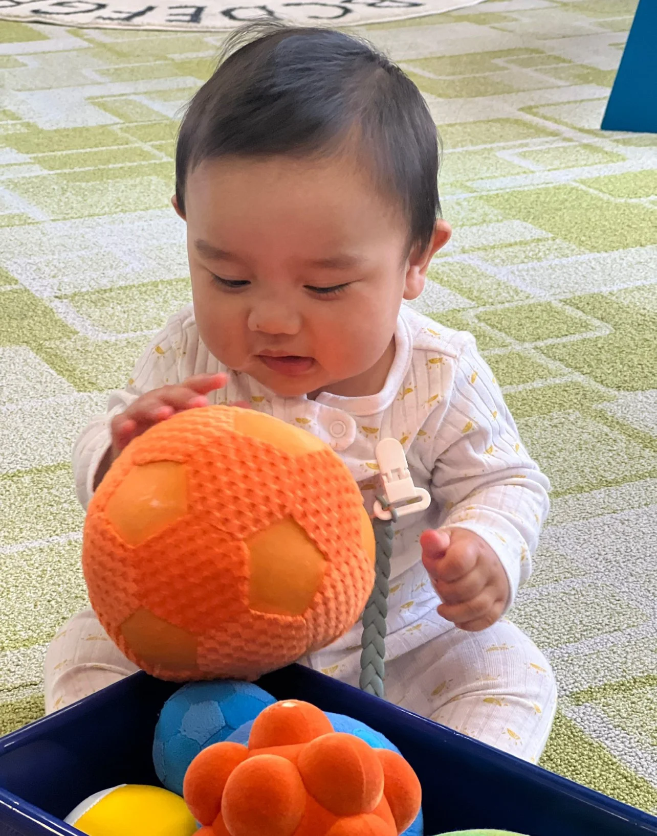 A young child with dark hair sitting on a green and white patterned carpet, holding an orange textured ball, and looking at it with curiosity, with a box of colorful soft balls in front.