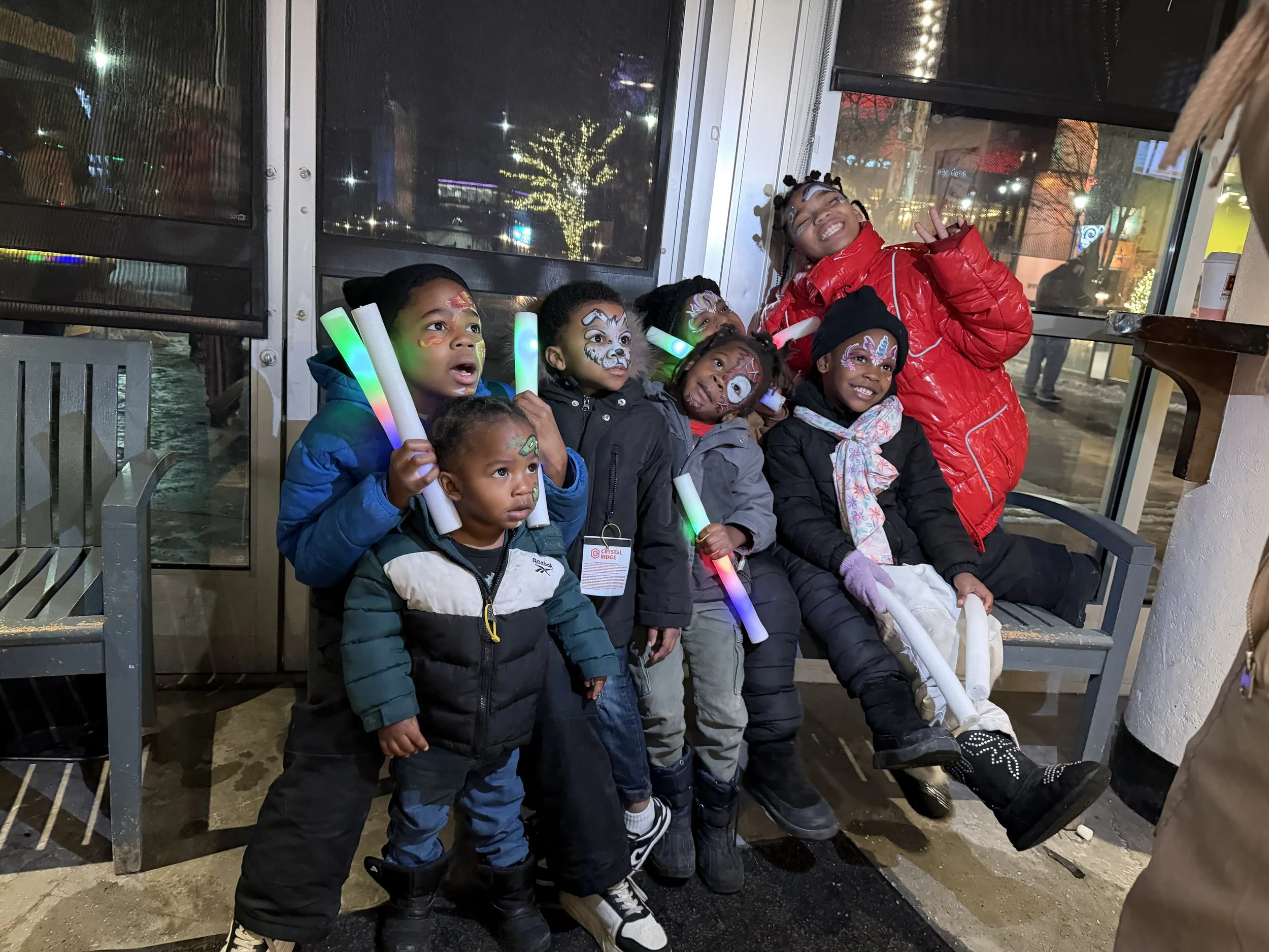 Group of six children sitting on a bench indoors, dressed in warm clothing, with face paint and holding glowing light sticks, and an adult woman standing behind them smiling and making a peace sign, nighttime outside visible through the windows.