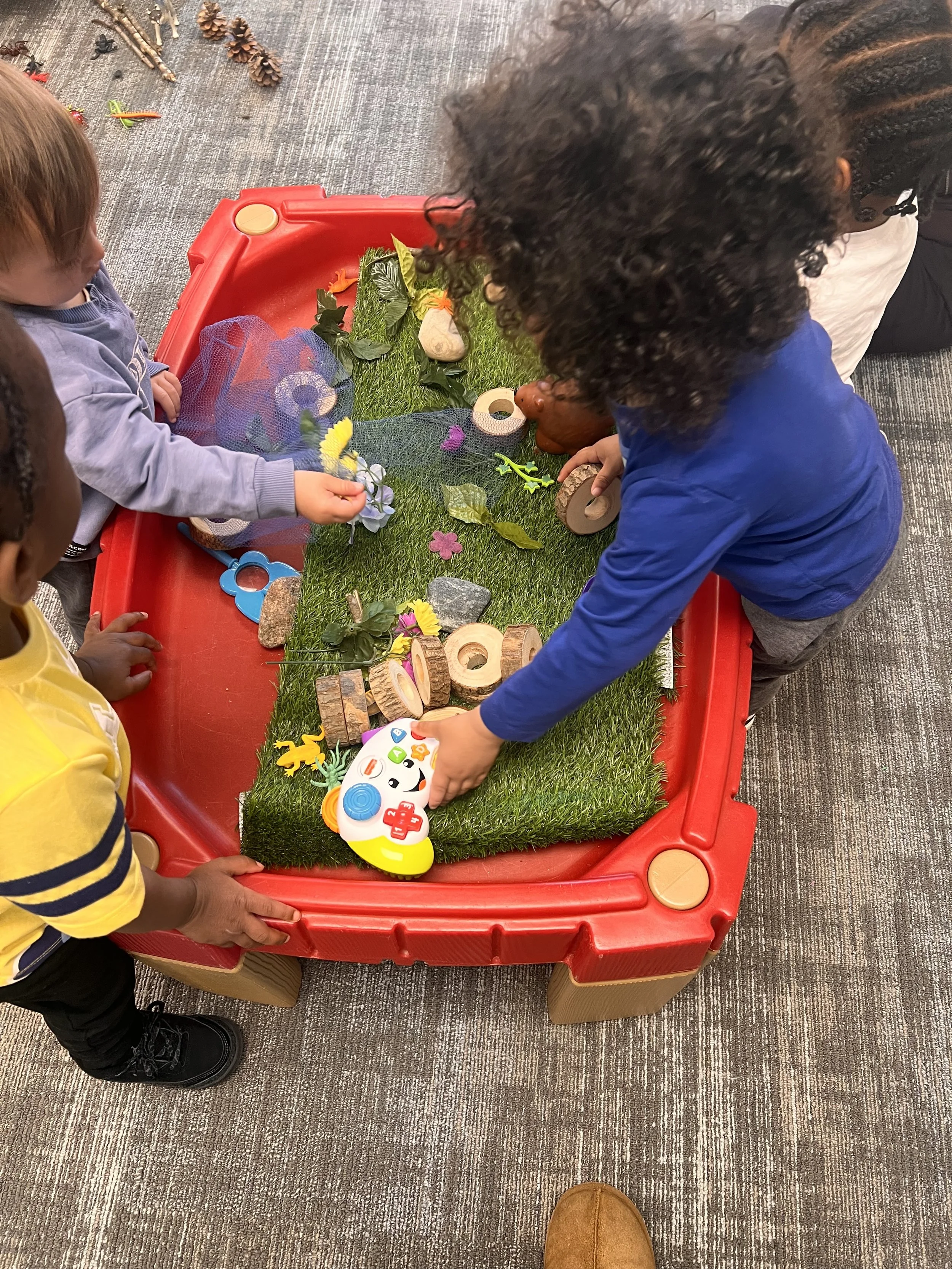 Children playing with a sensory bin filled with artificial grass, wooden blocks, leaves, flowers, and toy animals.