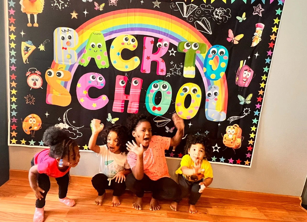 Four children, three girls and one boy, are sitting and kneeling on a wooden floor in front of a black birthday banner with colorful letters spelling "Birthday School" and illustrated decorations. The children appear excited and are making playful gestures.