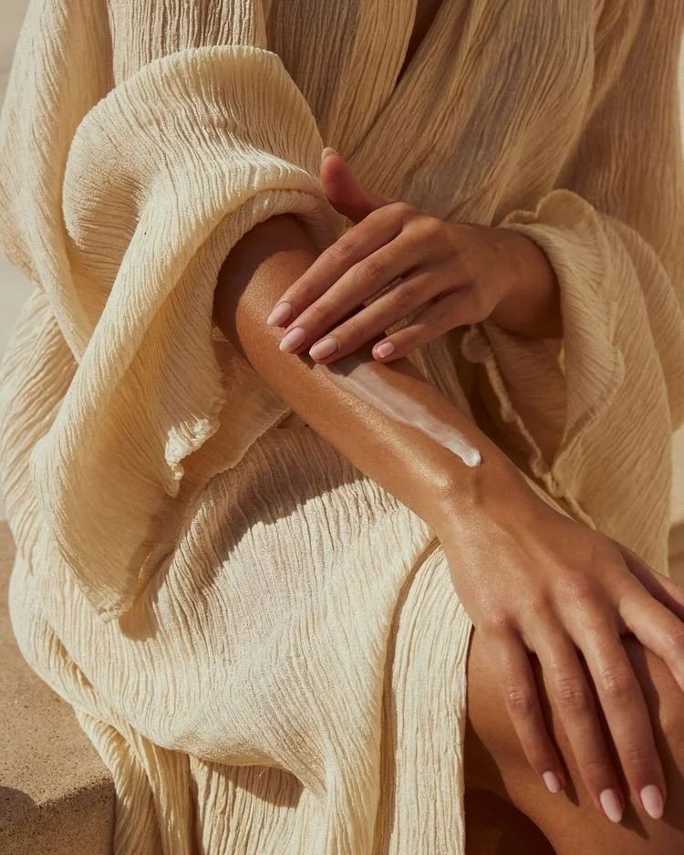 Person in a flowy beige dress applying sunscreen to their arm.