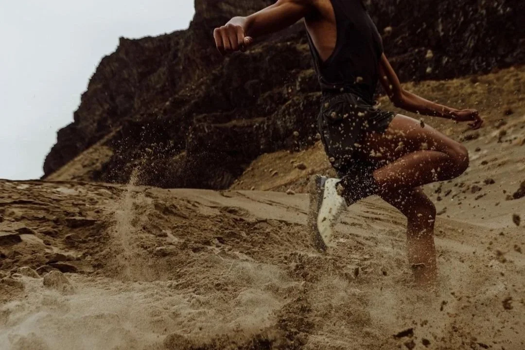 Person running or jumping on sandy terrain with rocky cliffs in the background.