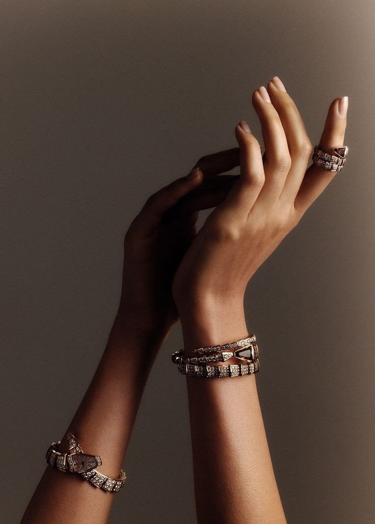 A pair of hands, one with a ring and multiple bracelets, reaching upwards against a plain background.