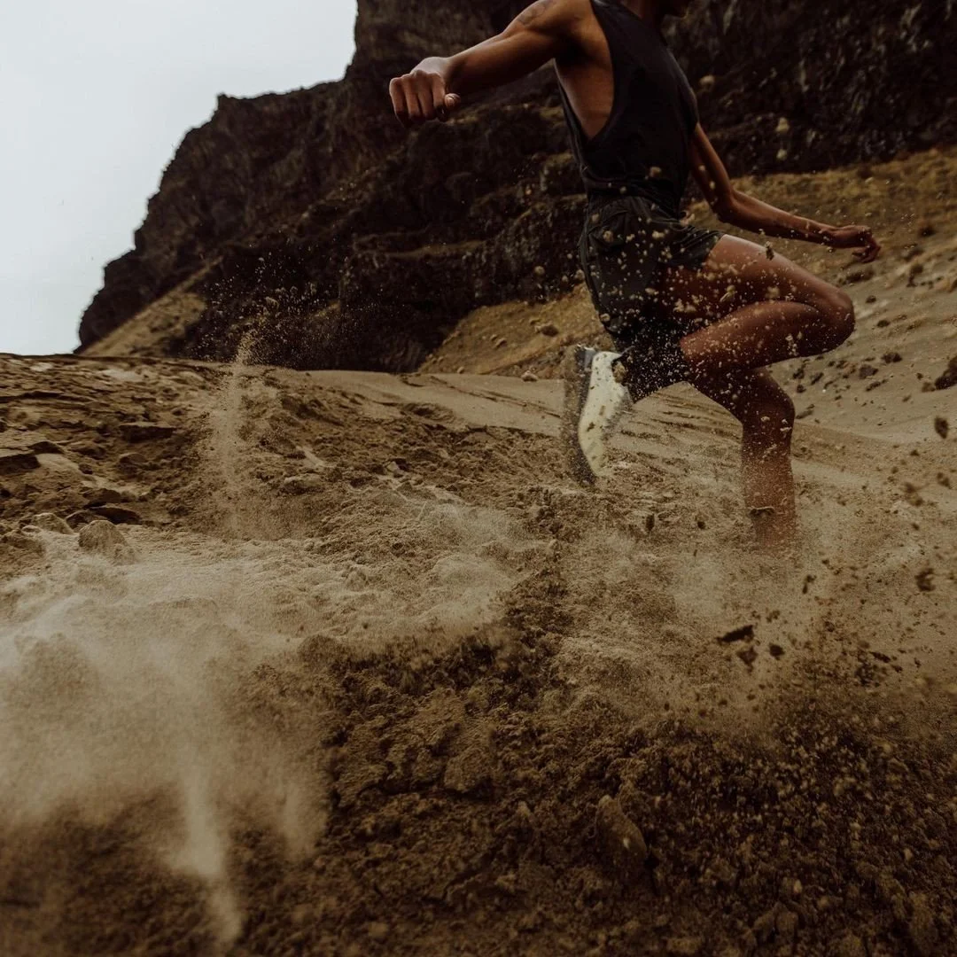 A person wearing a black tank top and shorts running in sandy terrain outdoors, kicking up dust and sand.
