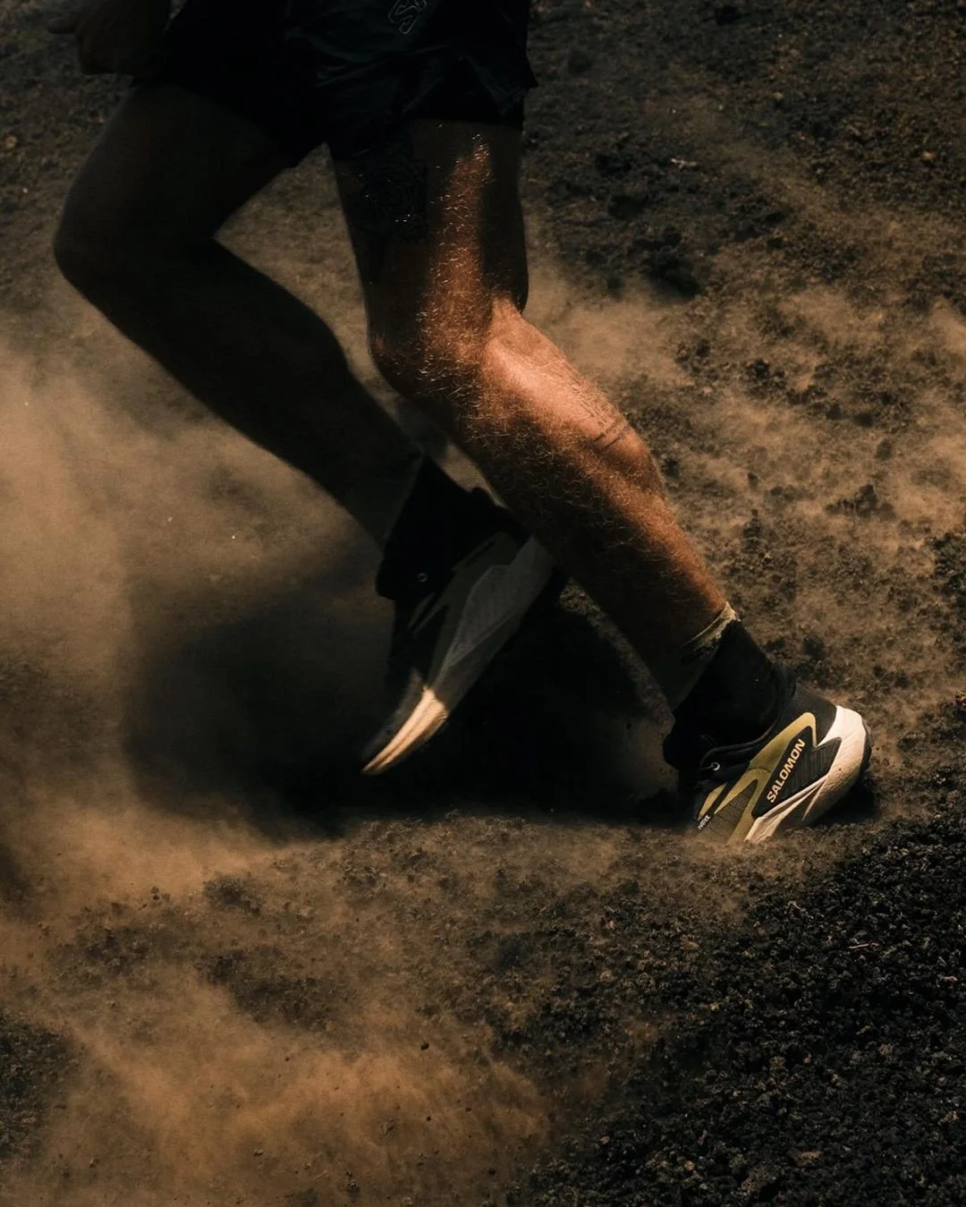 A person wearing black athletic shoes with Salomon branding, black pants, and a black jacket is falling or tumbling on a dirt or gravel surface, kicking up dust.