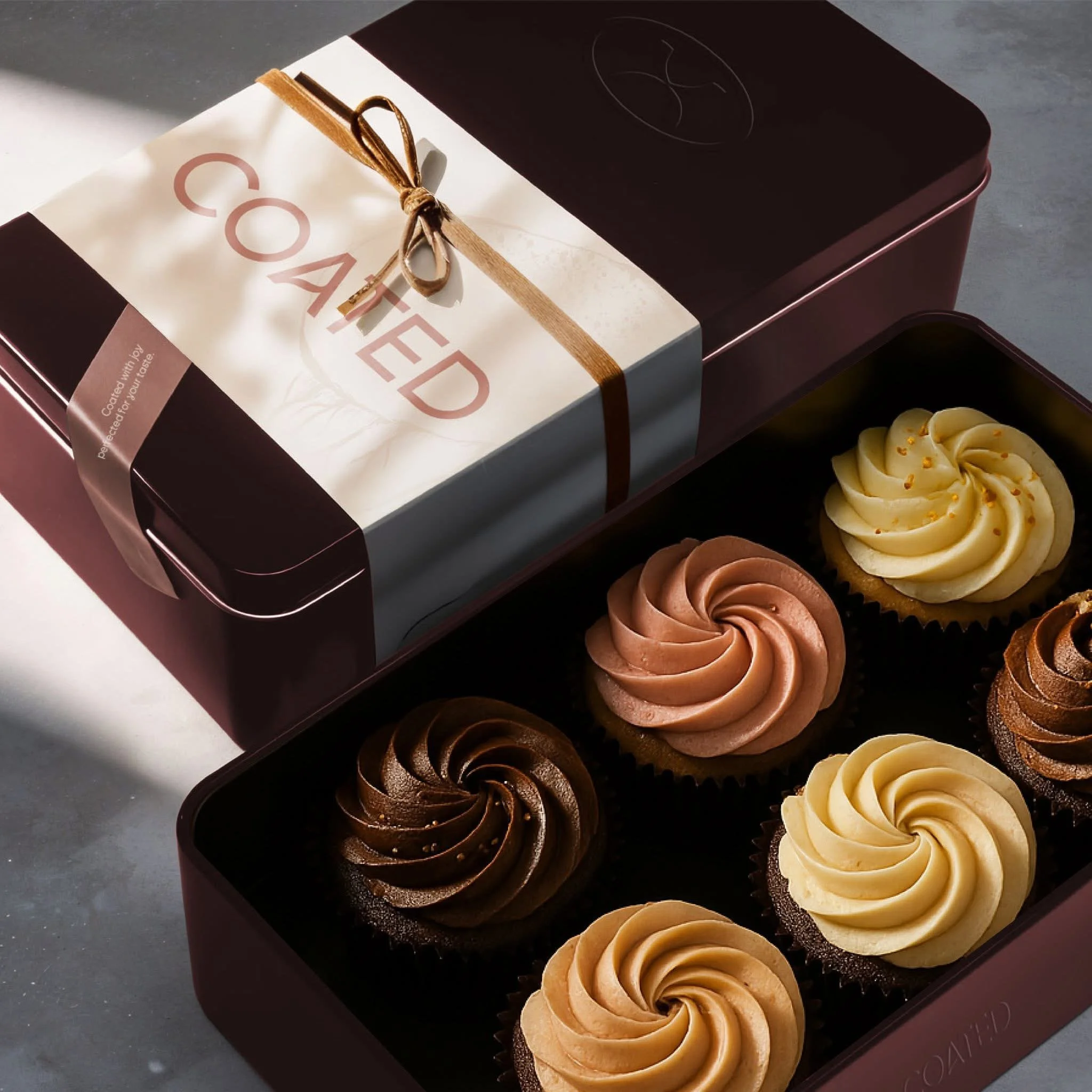 A box of assorted cupcakes with swirls of frosting in different colors and decorative sprinkles, placed on a dark surface.