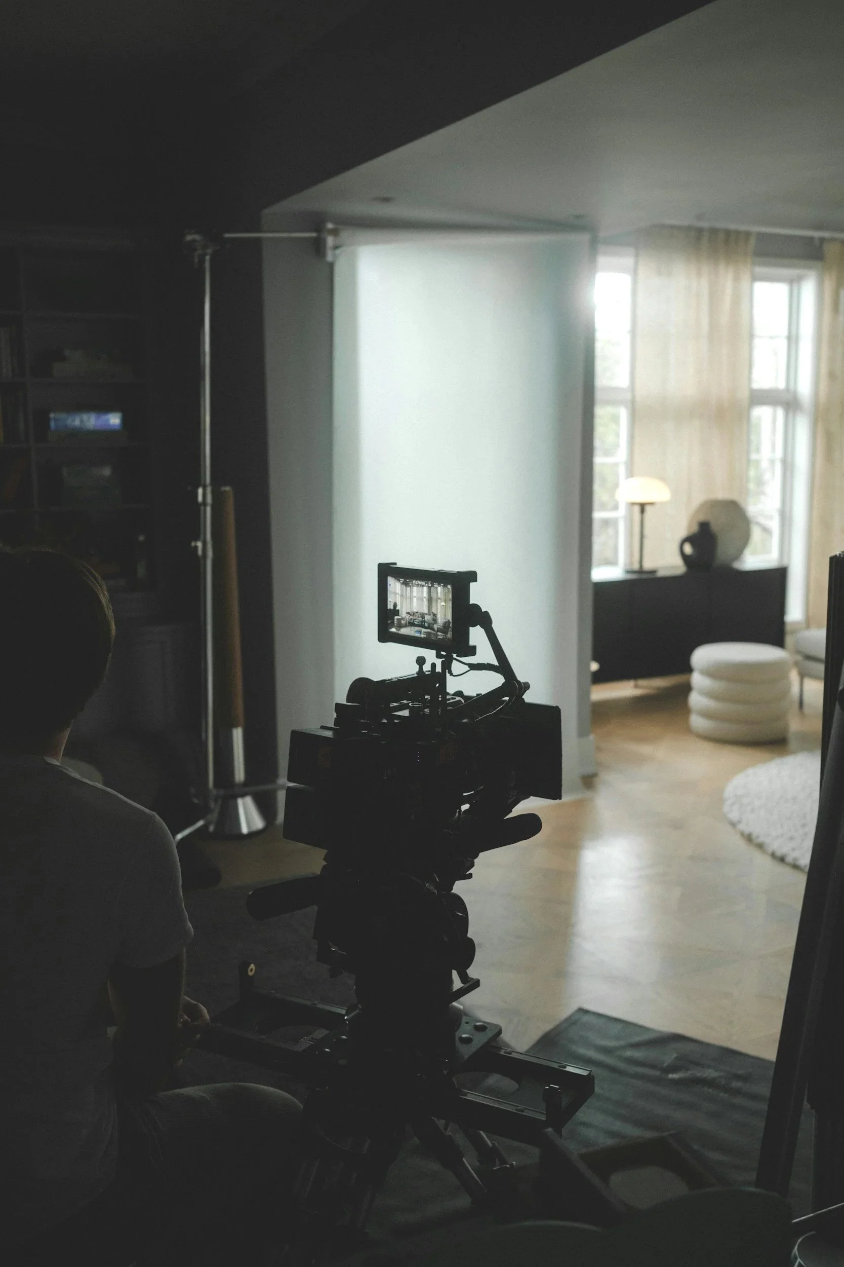 Film or photography studio setup with camera on track, a person in the foreground, lighting equipment, and a living room background with curtains, a lamp, and decorative vases.