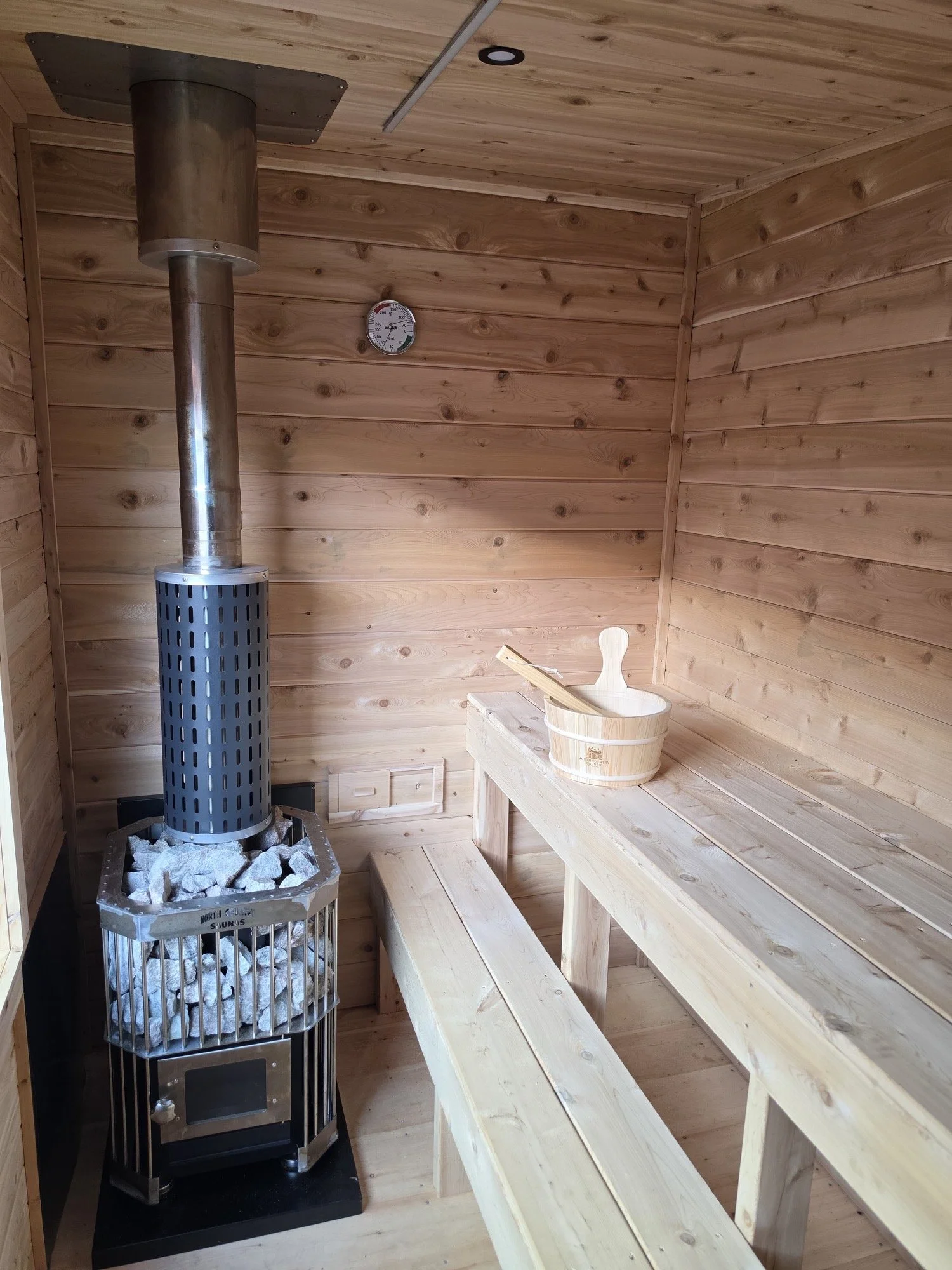 Wood-paneled sauna room with a stove filled with rocks, a wood bucket, and benches.