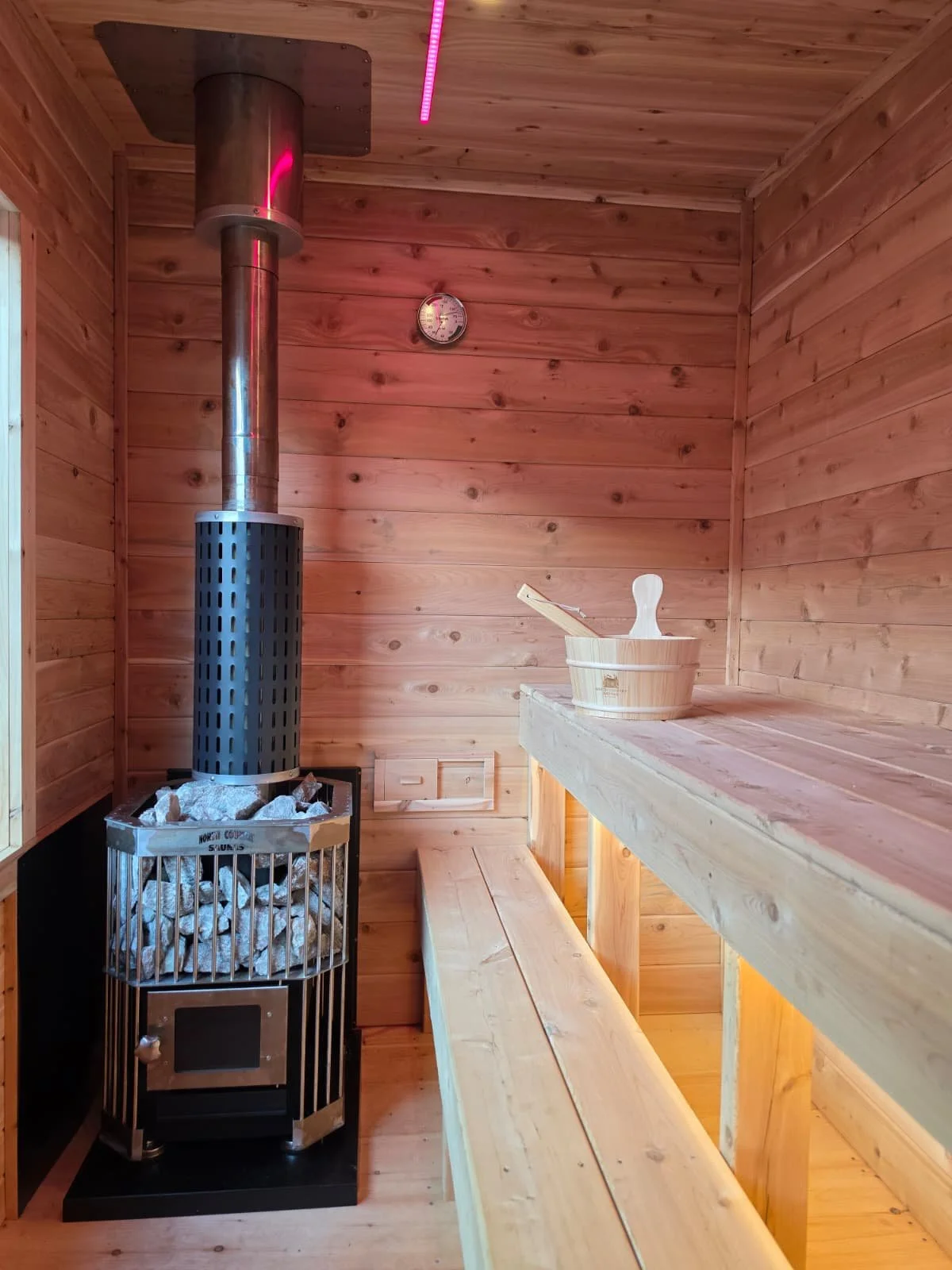 Wood-paneled sauna room featuring a metal stove with rocks on top, a wooden bucket with a ladle, and a bench along the wall.