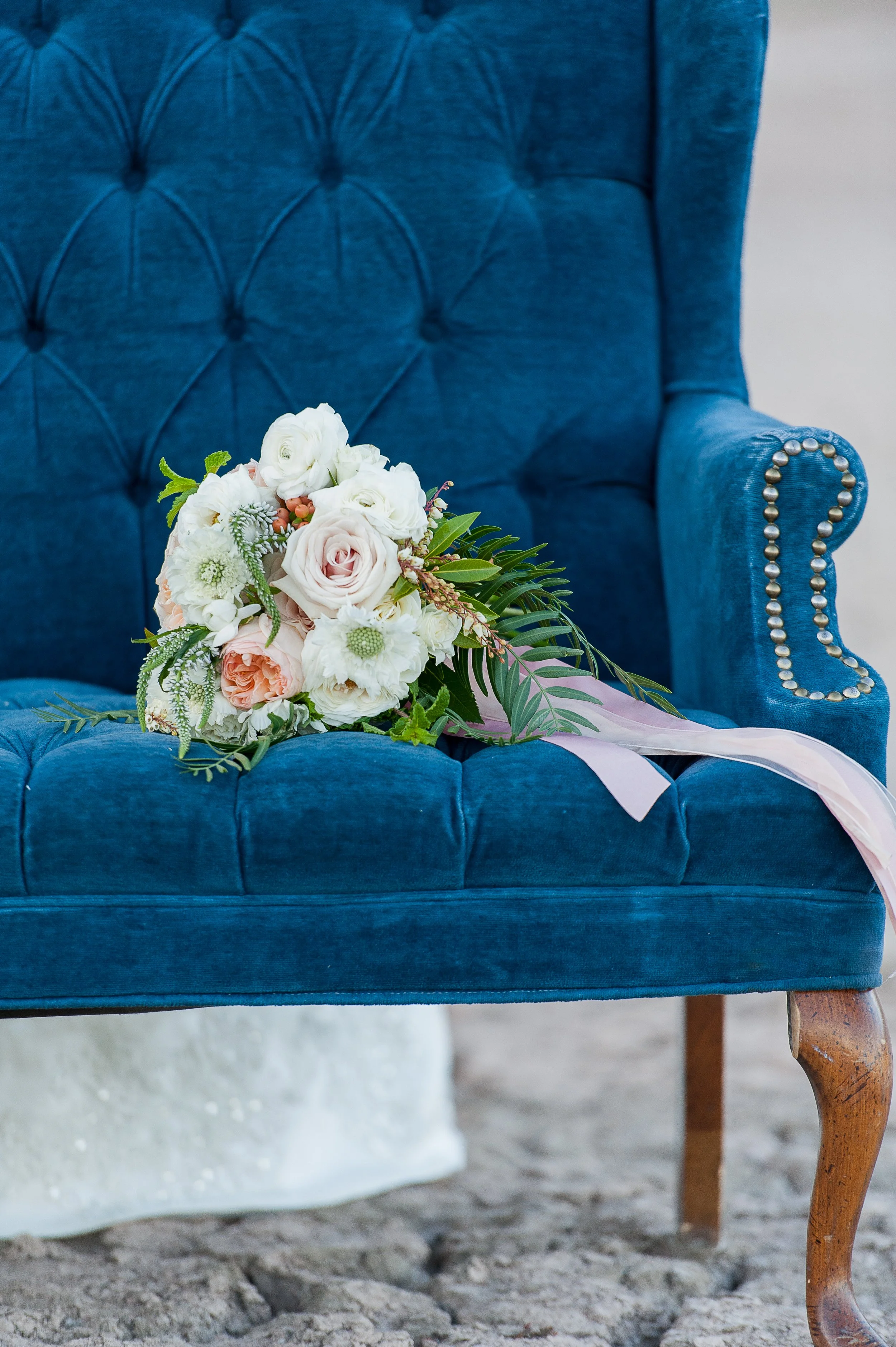 A bouquet of flowers resting on the seat of a blue velvet armchair with a wooden leg, placed on a cobblestone surface.