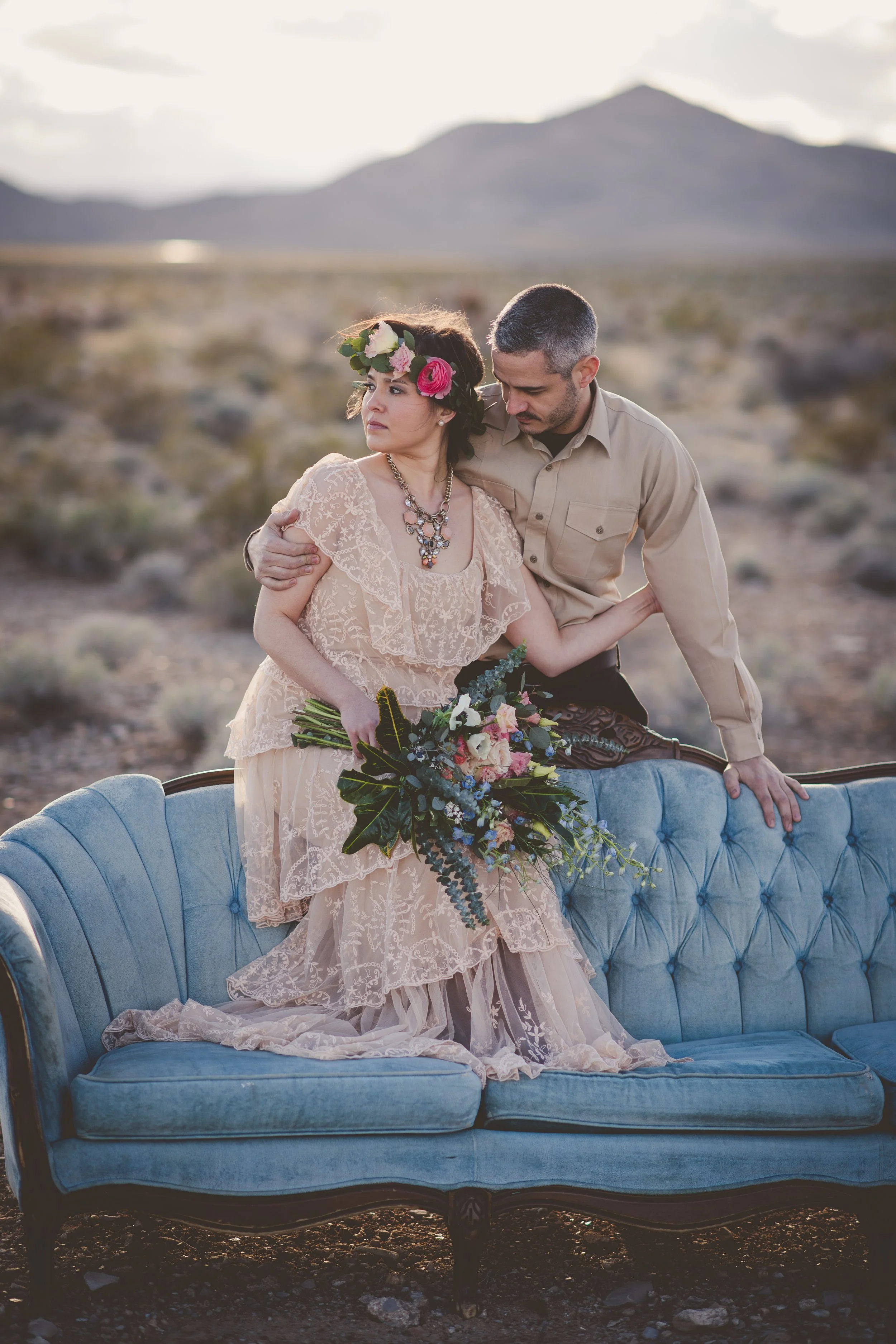 A woman in a lace wedding dress with a floral crown and holding a bouquet, sitting on a vintage blue velvet sofa in a desert landscape with a man in a beige shirt behind her.