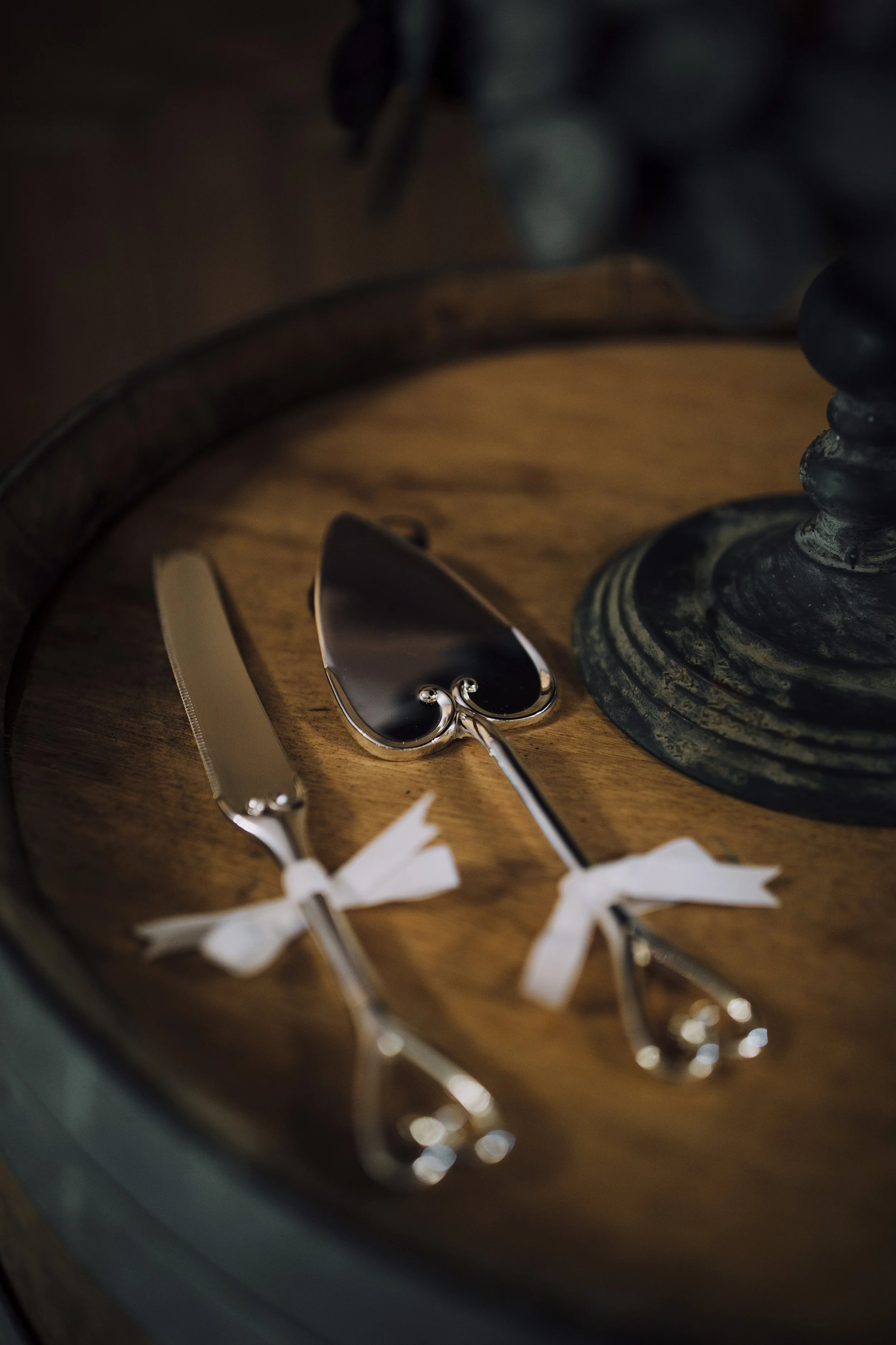 Close-up of a wooden tray holding a pair of silver scissors with white ribbon tied around the handles, a nail file, and a round decorative object with a black and white pattern, partially visible. The scene appears dimly lit.
