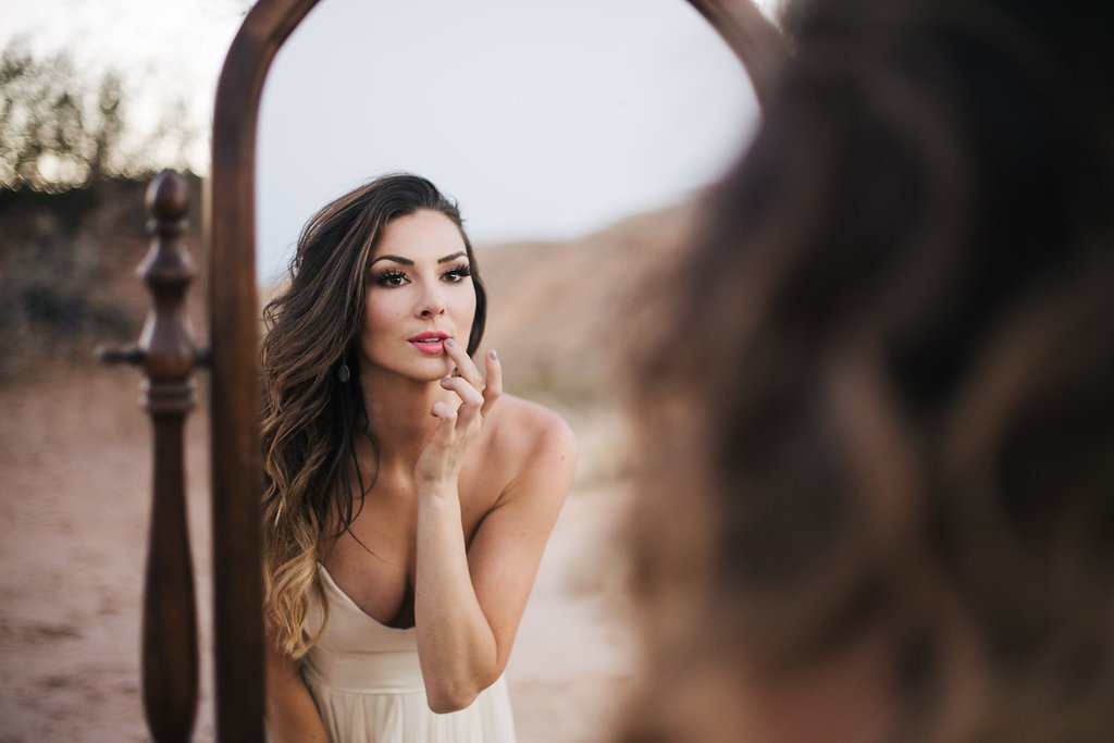 A woman looking into a mirror with a thoughtful expression, touching her lips with her finger, outdoors with a blurred natural background.