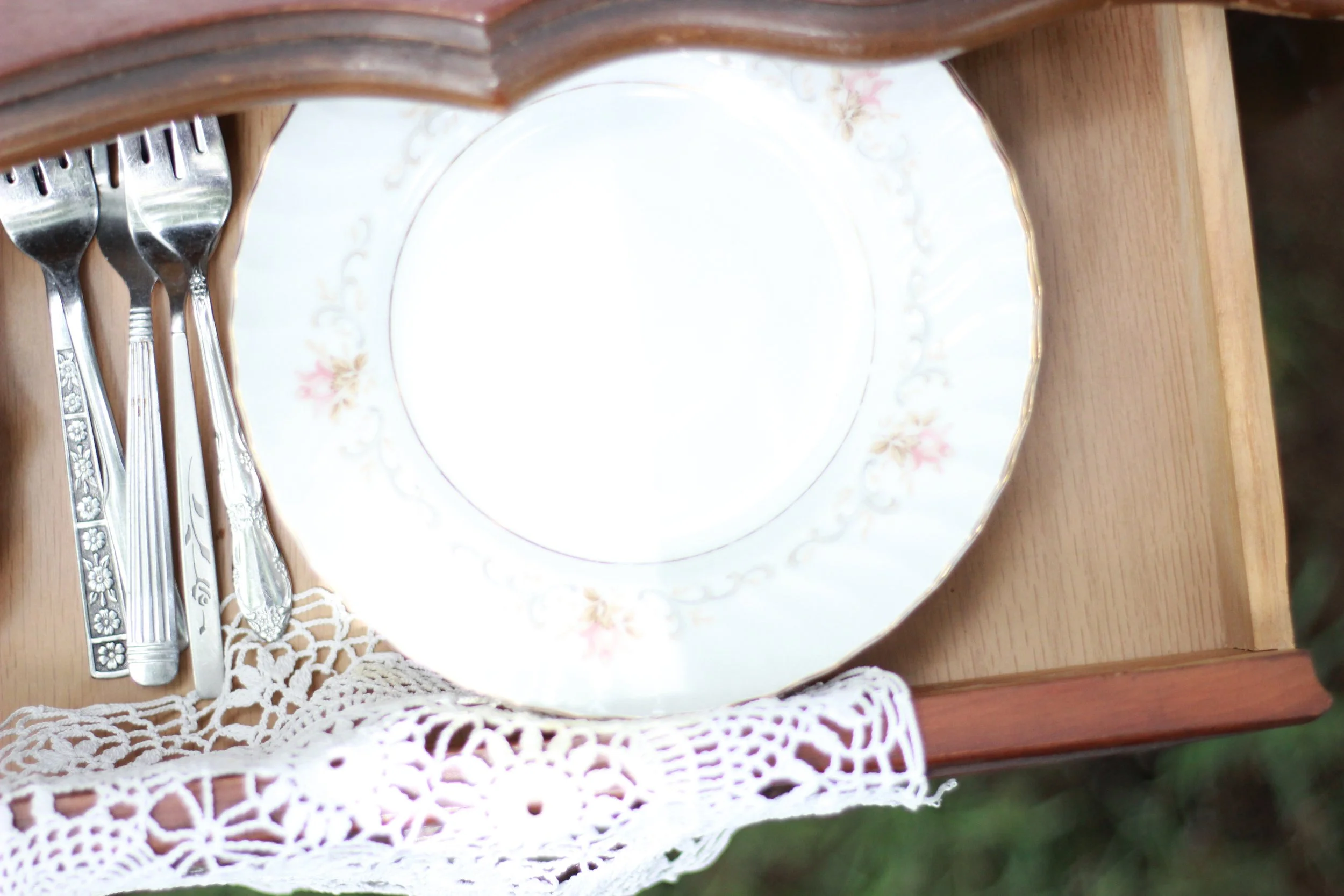 A wooden table set with four silver forks on the left, a decorative porcelain plate with floral patterns in the center, and white crocheted doilies underneath the plate and forks.