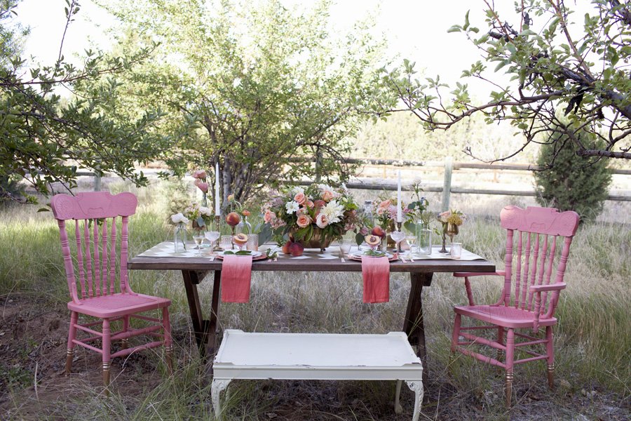 An outdoor dining setup with a wooden table decorated with pink and white flowers, pink chairs, and a white bench, under trees in a grassy yard.