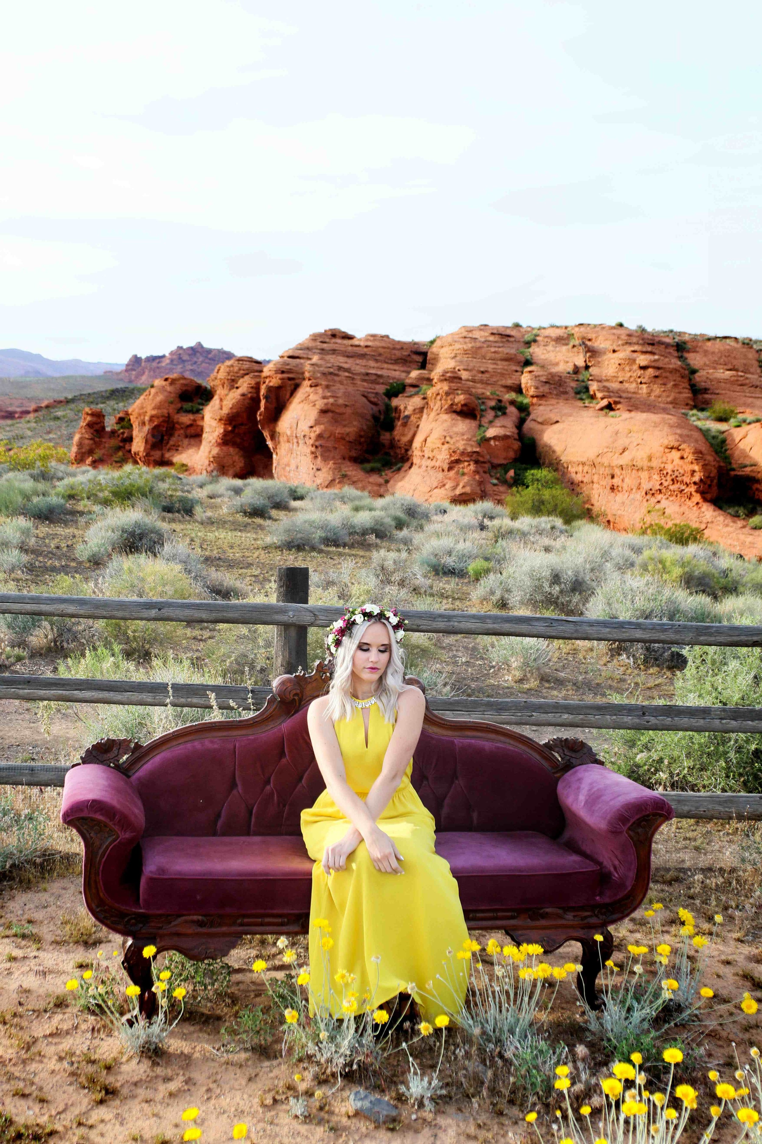A woman with long blonde hair wearing a yellow dress and a flower crown, sitting on a vintage velvet sofa outdoors with a desert landscape and red rock formations in the background.