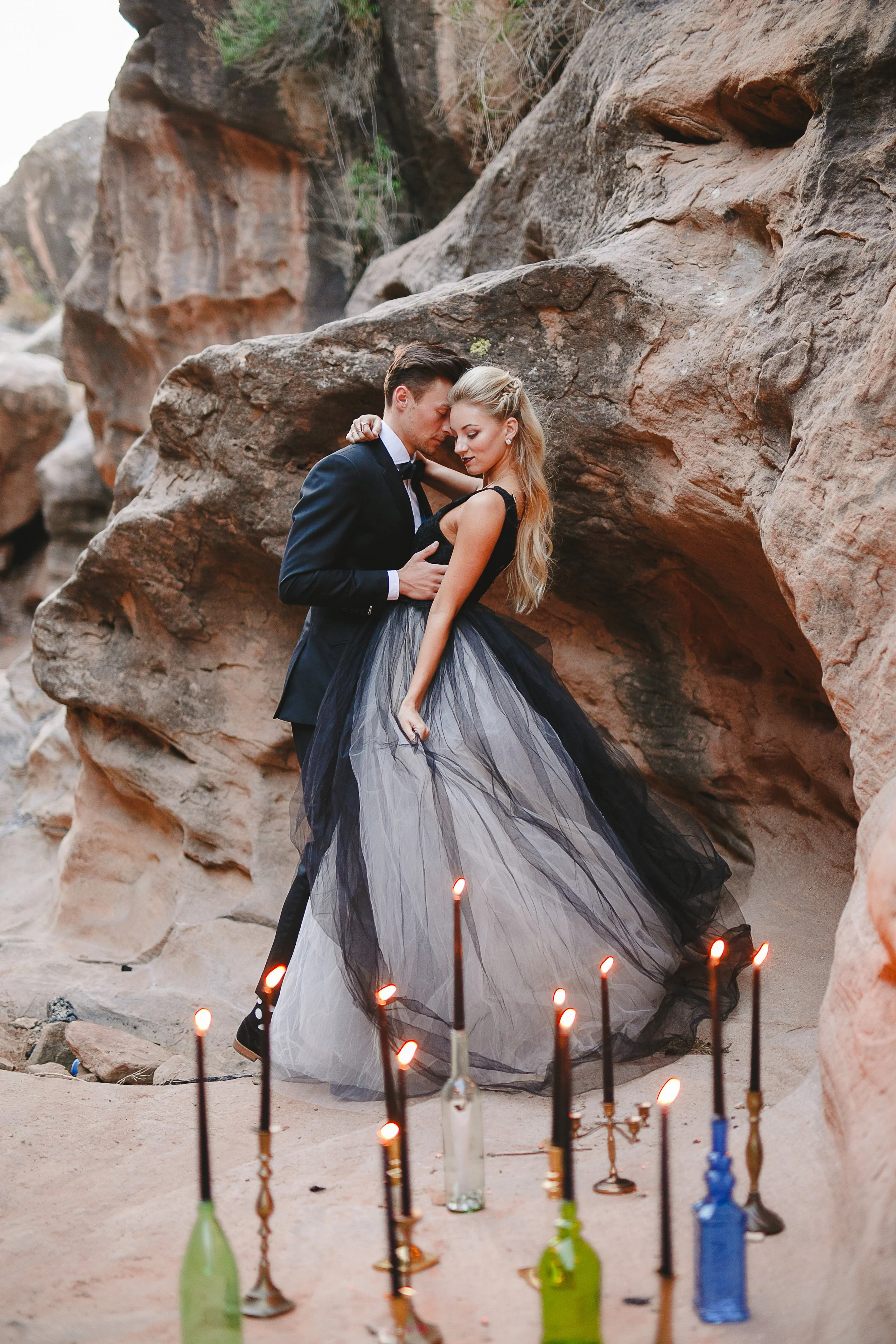 A couple dressed in formal black attire, with the woman in a flowing black and gray gown, embracing and touching foreheads in front of a rocky outdoor setting. There are lit candles in glass bottles on the ground in front of them, with a backdrop of large rugged rocks.