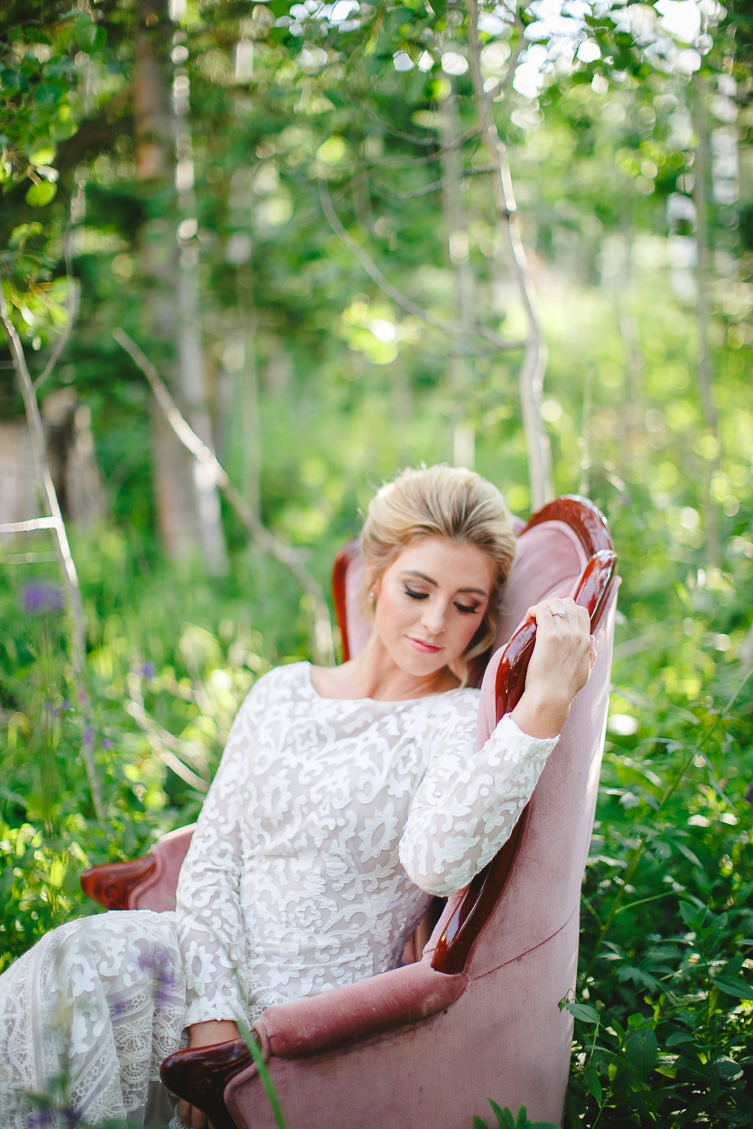A woman with blonde hair in a white lace dress sitting on a pink velvet armchair outdoors in a garden surrounded by green plants and trees, looking relaxed with her eyes closed.