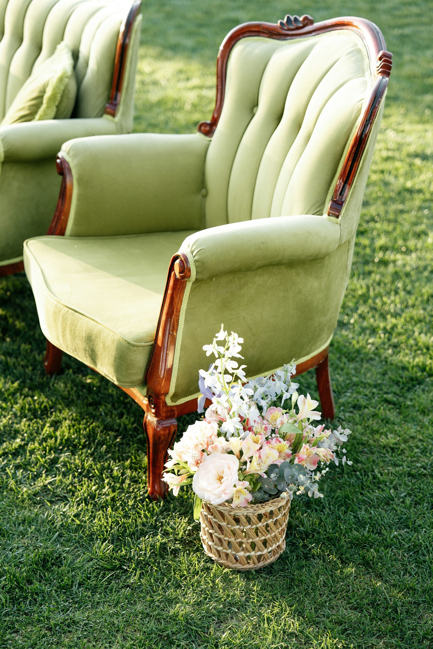 Green vintage armchair with wooden frame on a grassy lawn, surrounded by a basket of pink and white flowers.