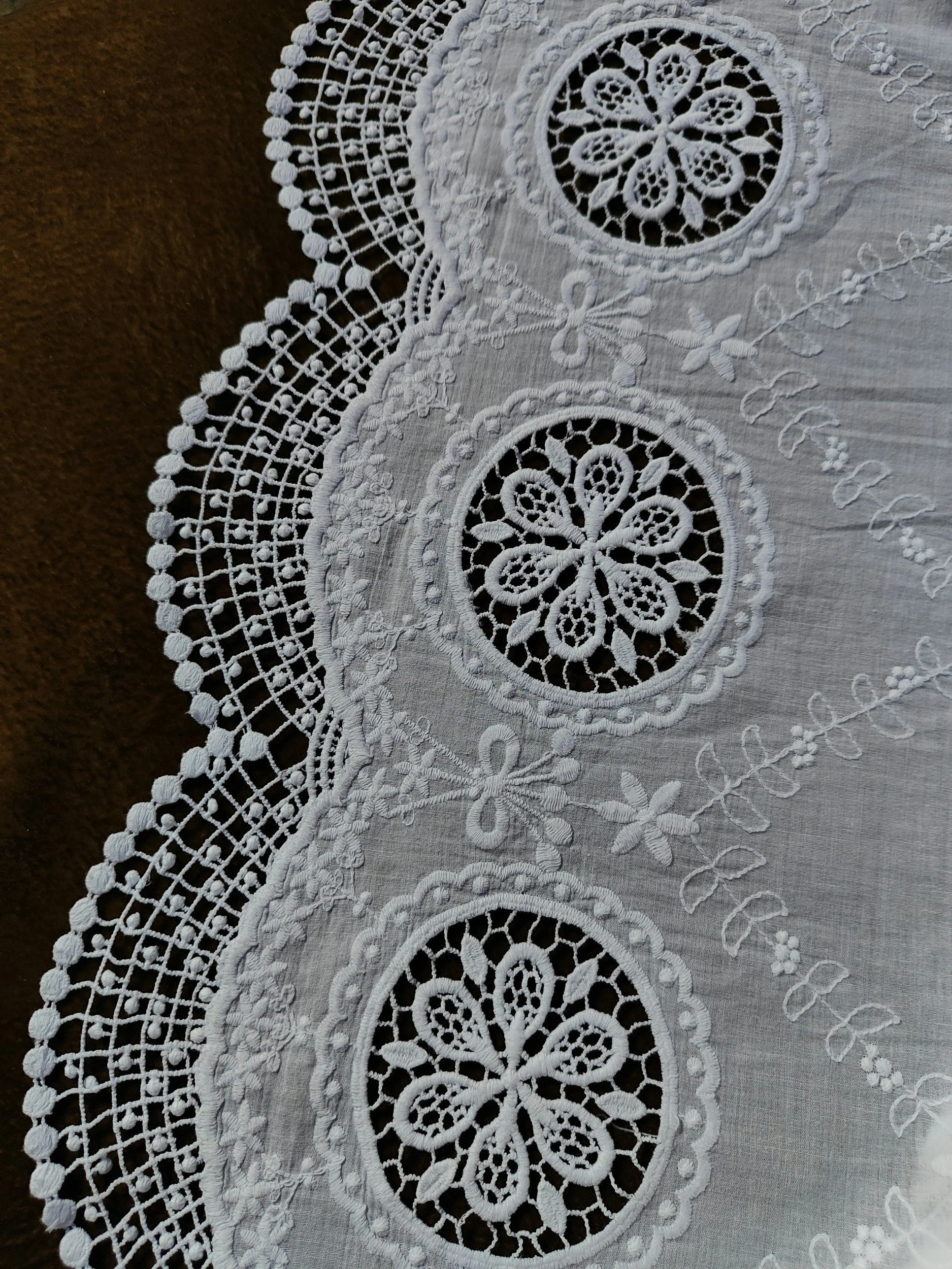 A close-up of a white embroidered lace tablecloth with intricate floral and geometric designs, including a pattern of large circular motifs and scalloped edges.