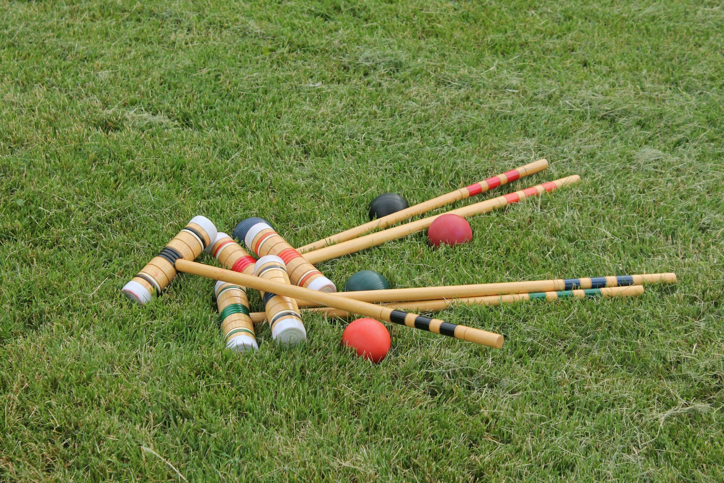 Croquet equipment scattered on a grassy lawn, including wooden mallets, plastic balls, and a wooden wicket.