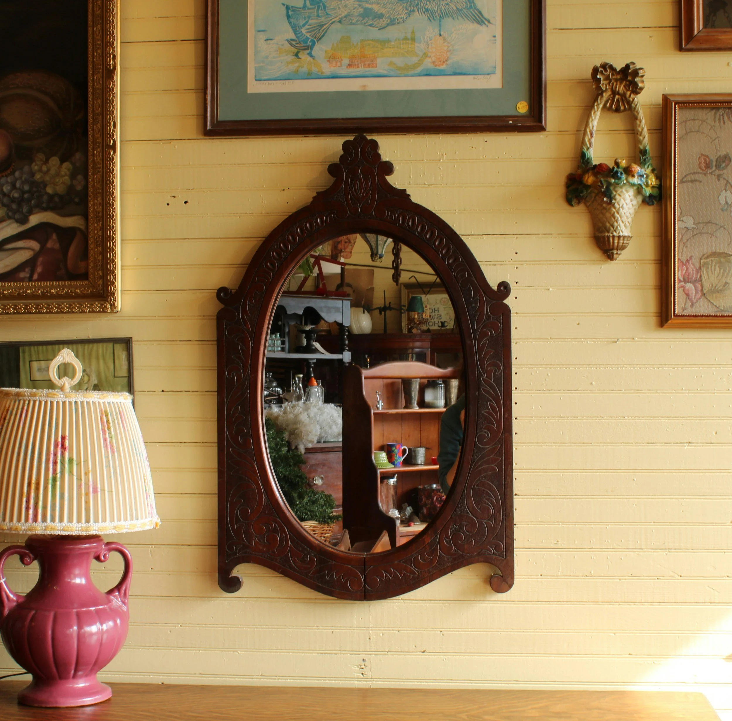 A wooden framed mirror with intricate carvings hangs on a yellow wooden wall. To the left of the mirror, there is a framed picture of grapes and a fish, and to the right, a decorative basket with artificial flowers. In front of the mirror, on a wooden surface, there is a pink ceramic vase and a lamp with a pleated fabric shade.