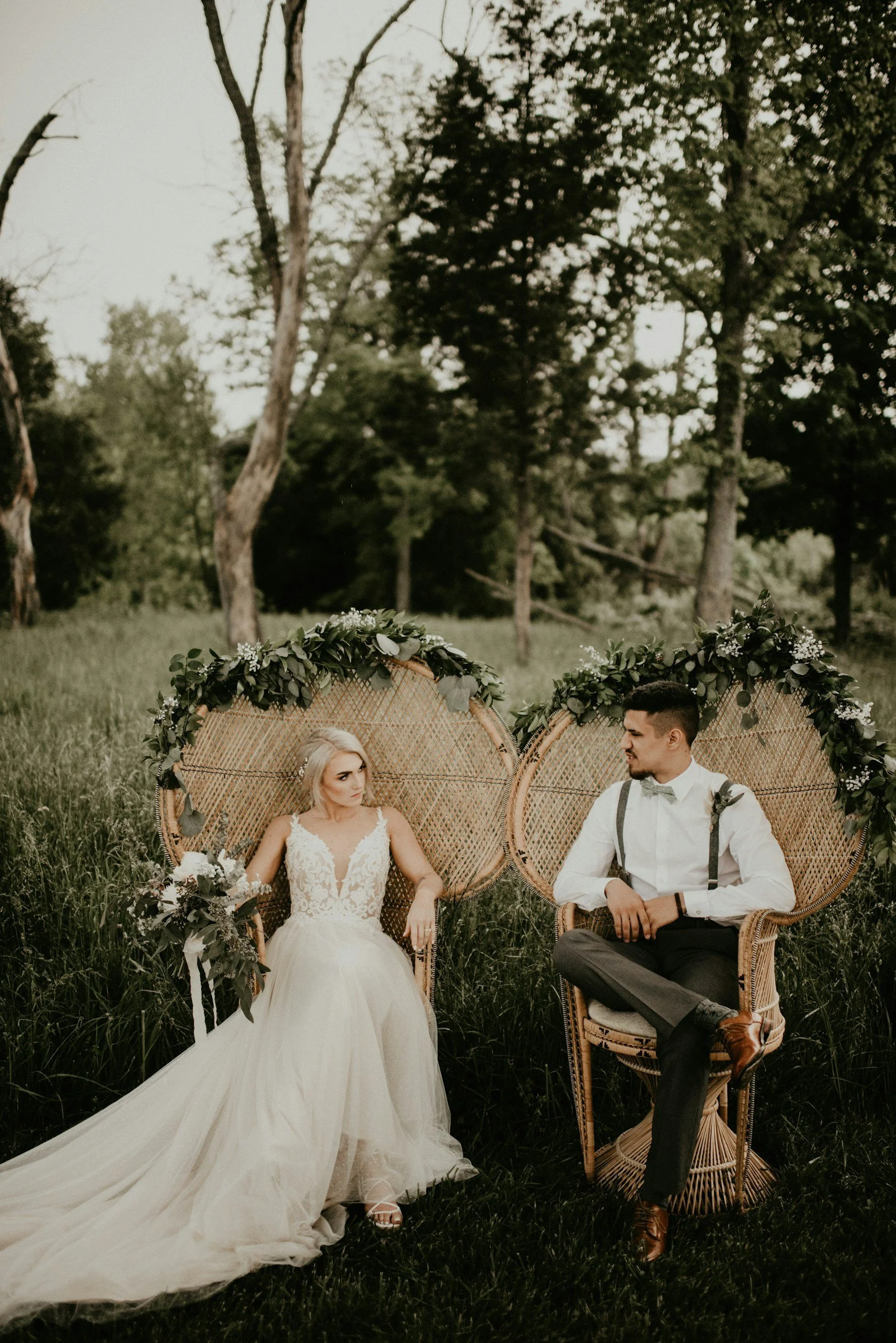 A bride and groom sitting in wicker chairs with greenery and floral decorations in an outdoor setting, surrounded by trees and grass.