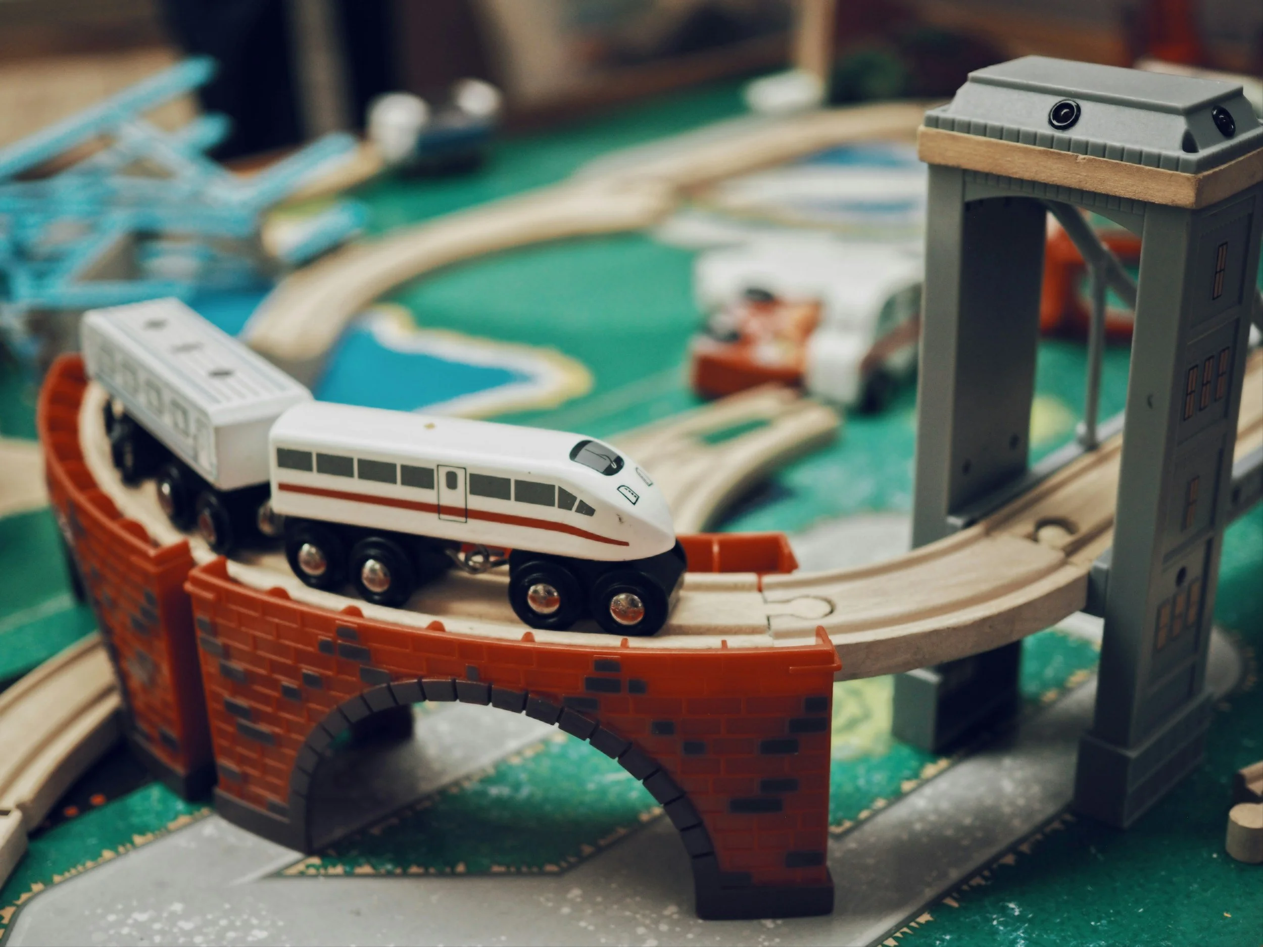 Close-up of a toy train on a model railway track, with a red twisted paper basket and a gray building structure in the background.