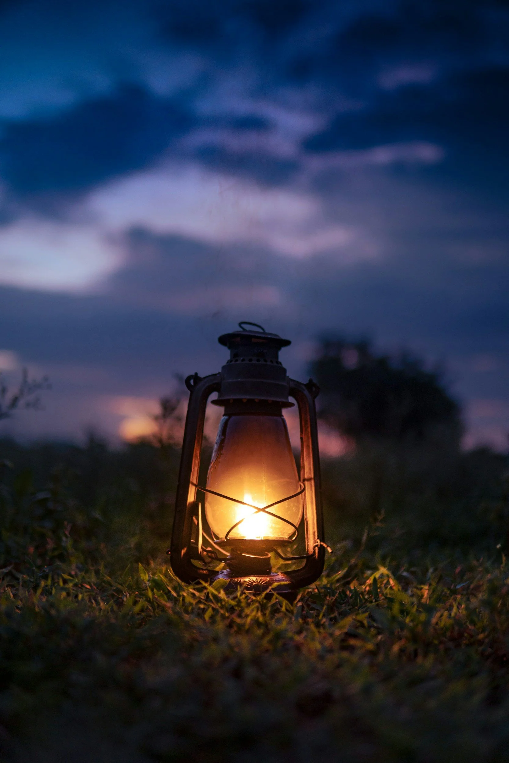 An old-fashioned lantern with a glowing flame, placed on grass outdoors during dusk or dawn with dark clouds in the sky.