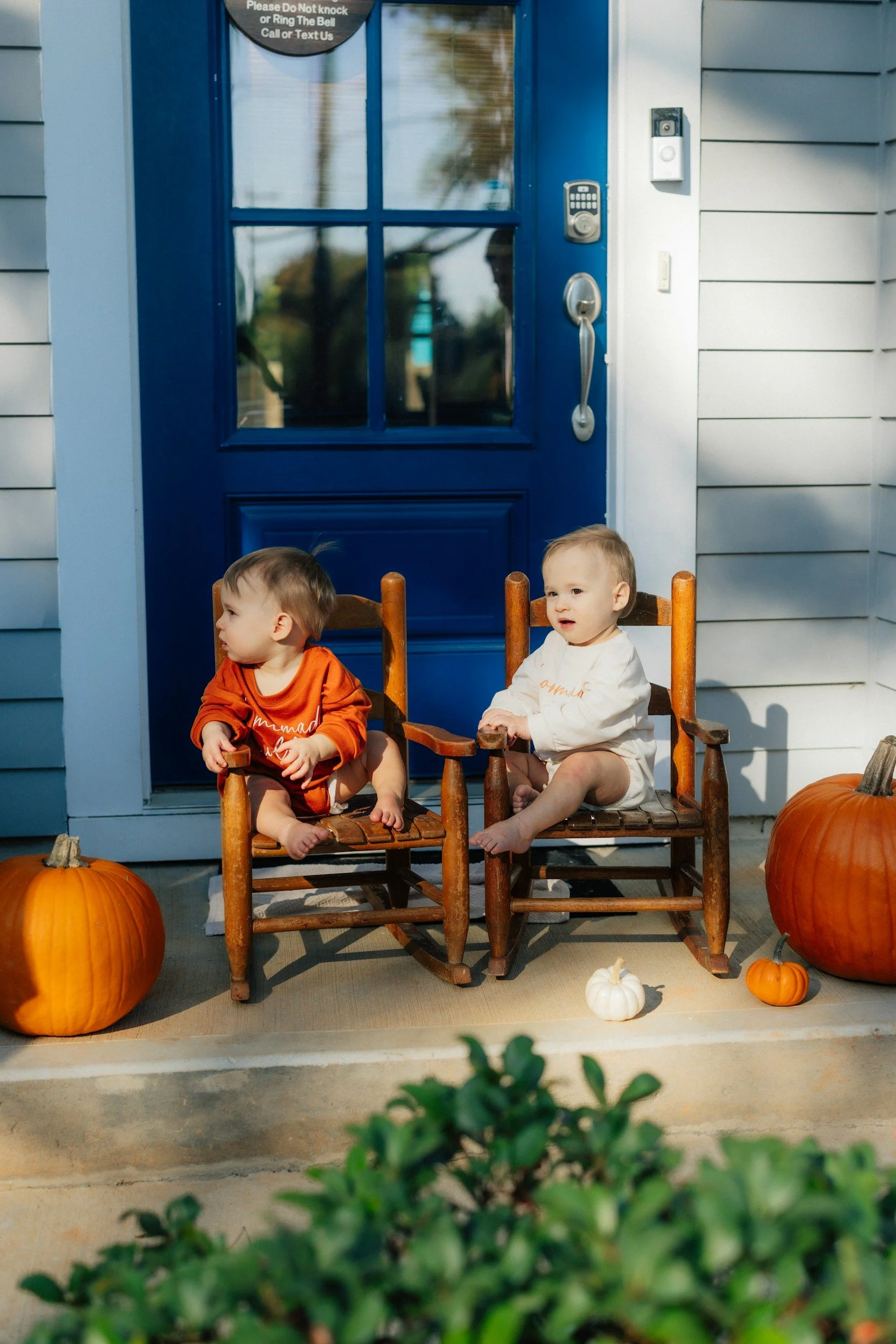 Two young children sitting in wooden chairs on a porch, surrounded by pumpkins, with a blue door and white siding behind them.